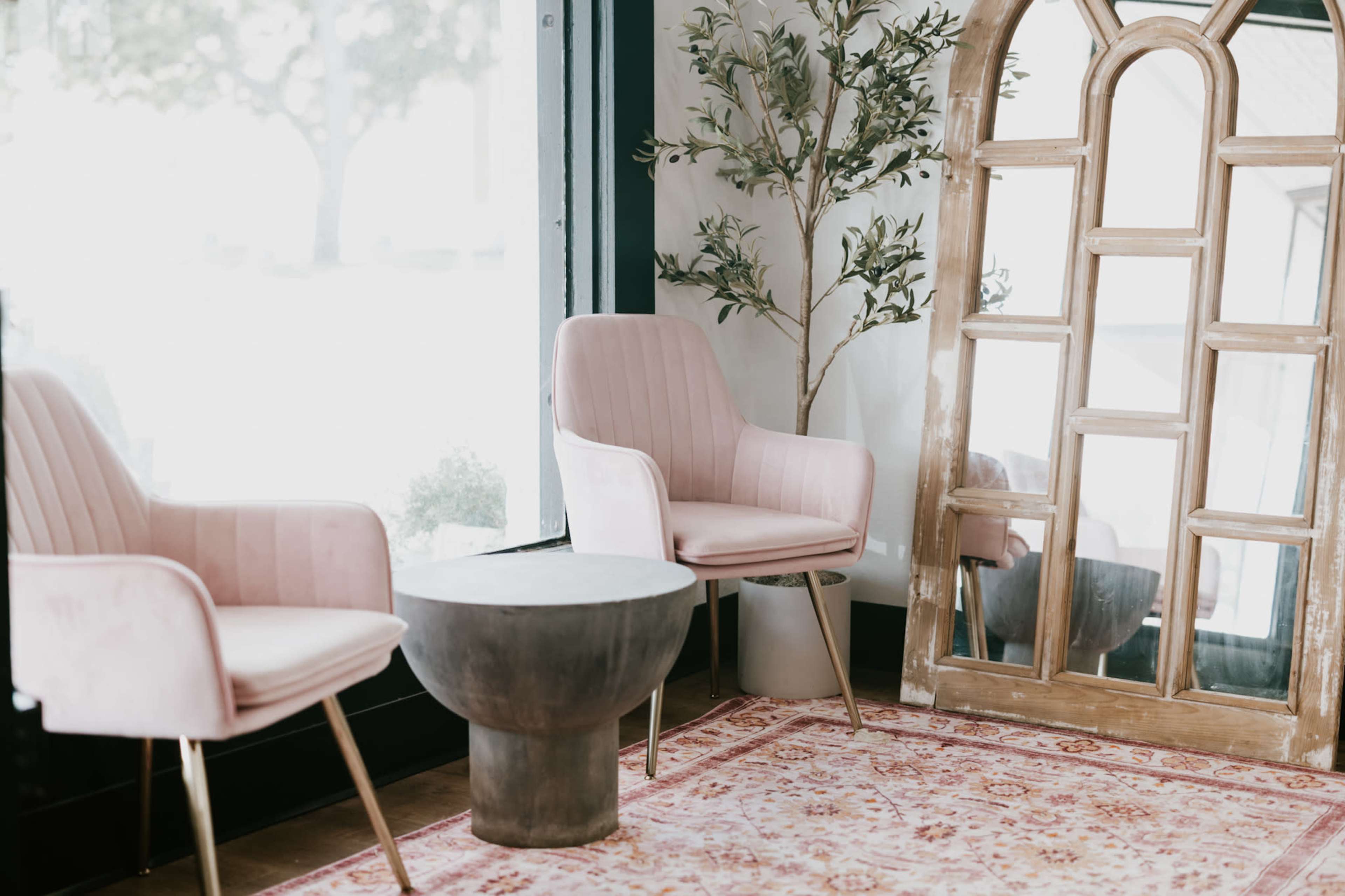 A cozy interior scene features two pink chairs, a round black table, and a tall wooden mirror beside a decorative indoor plant.