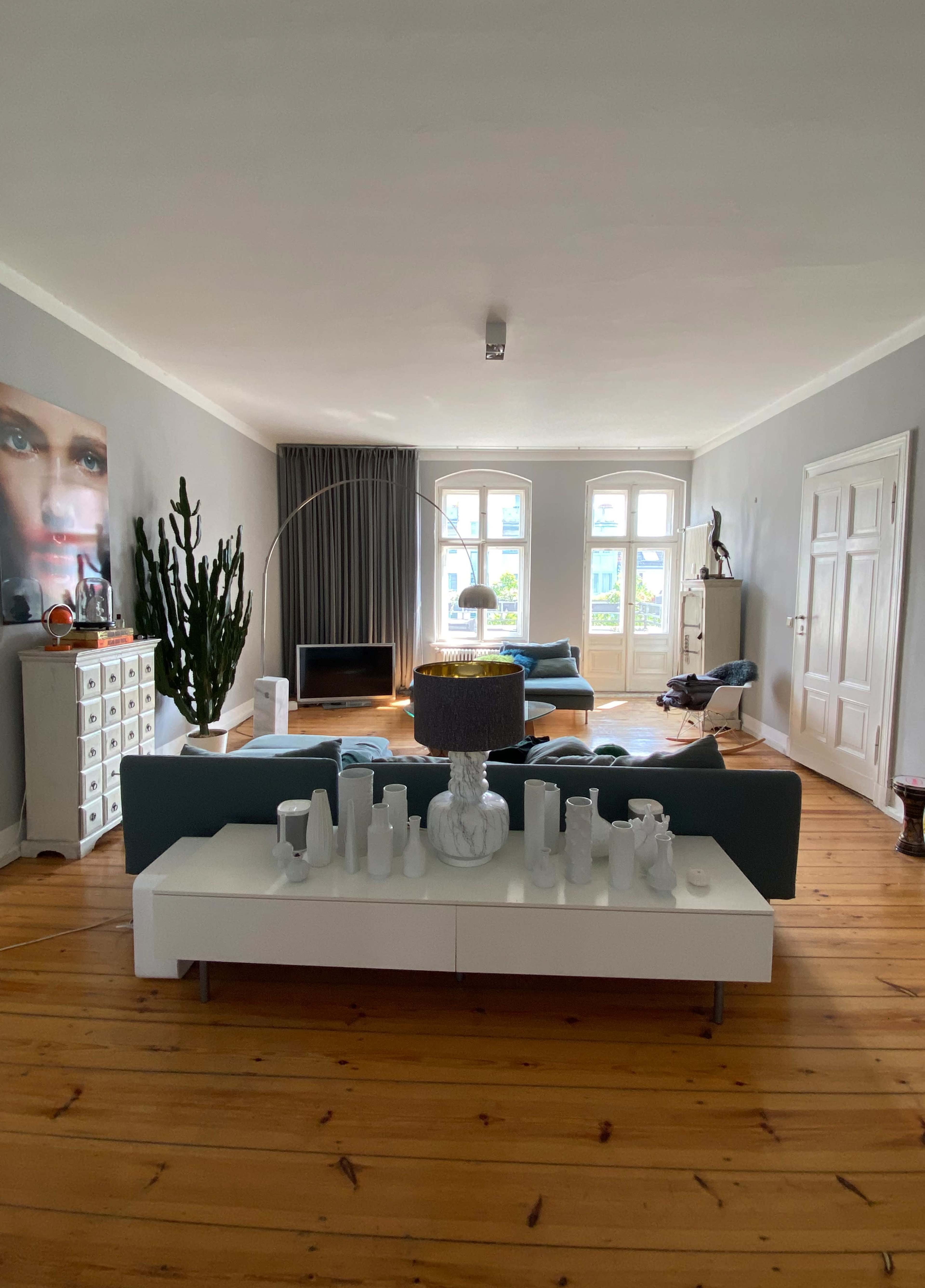 A spacious living room featuring green sofas, a white coffee table adorned with various vases, large windows with curtains, and a tall indoor plant.