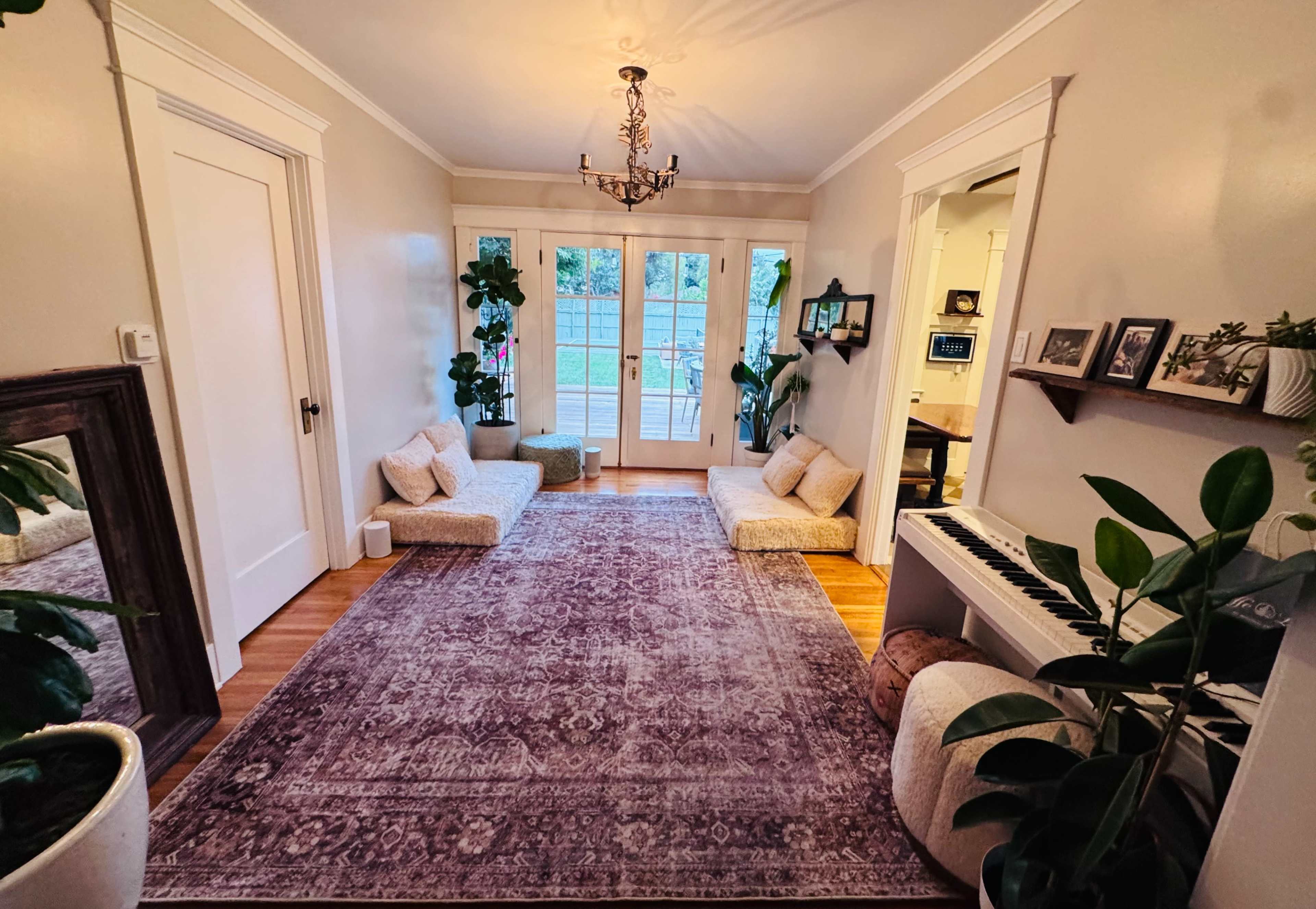 The image shows a cozy room with two seating areas by the windows, a decorative rug on the wooden floor, and a piano alongside various indoor plants.