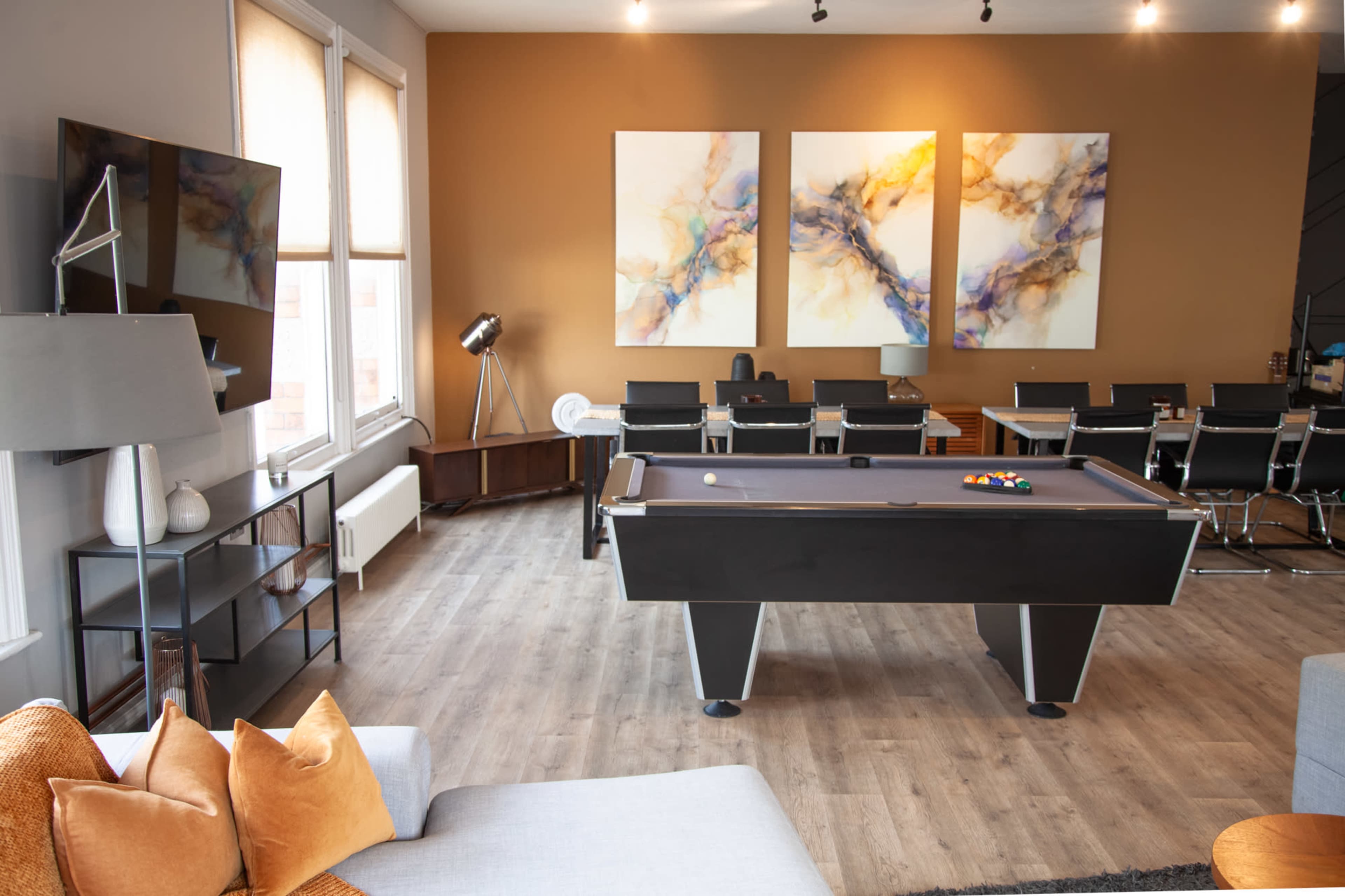 The image shows a modern game room featuring a pool table in the center, surrounded by a comfortable seating area and a dining space with sleek chairs and a stylish table.