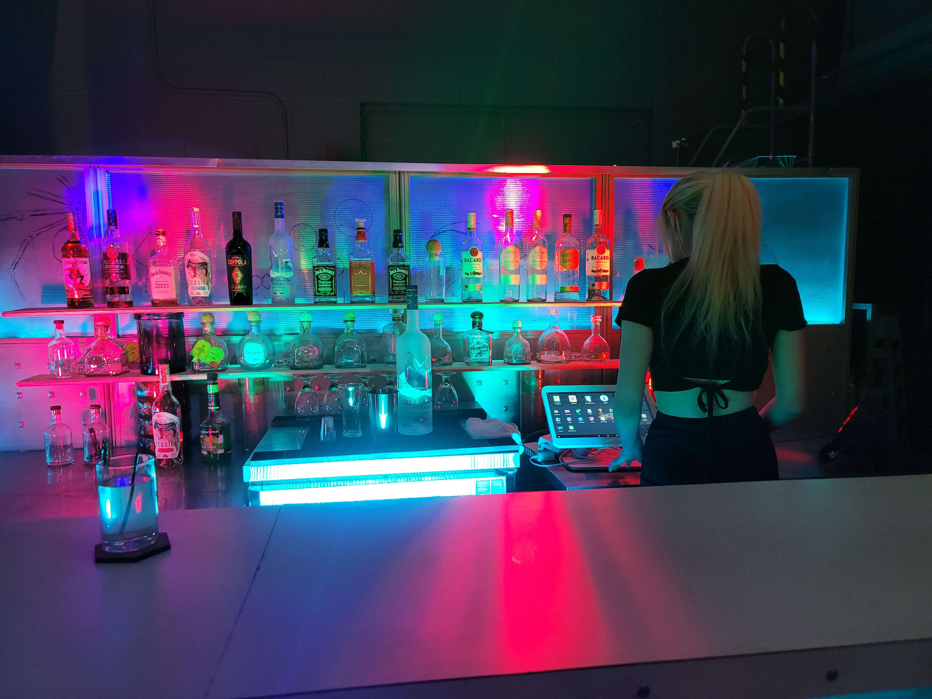 A bartender prepares drinks at a brightly lit bar featuring various bottles and illuminated shelves.