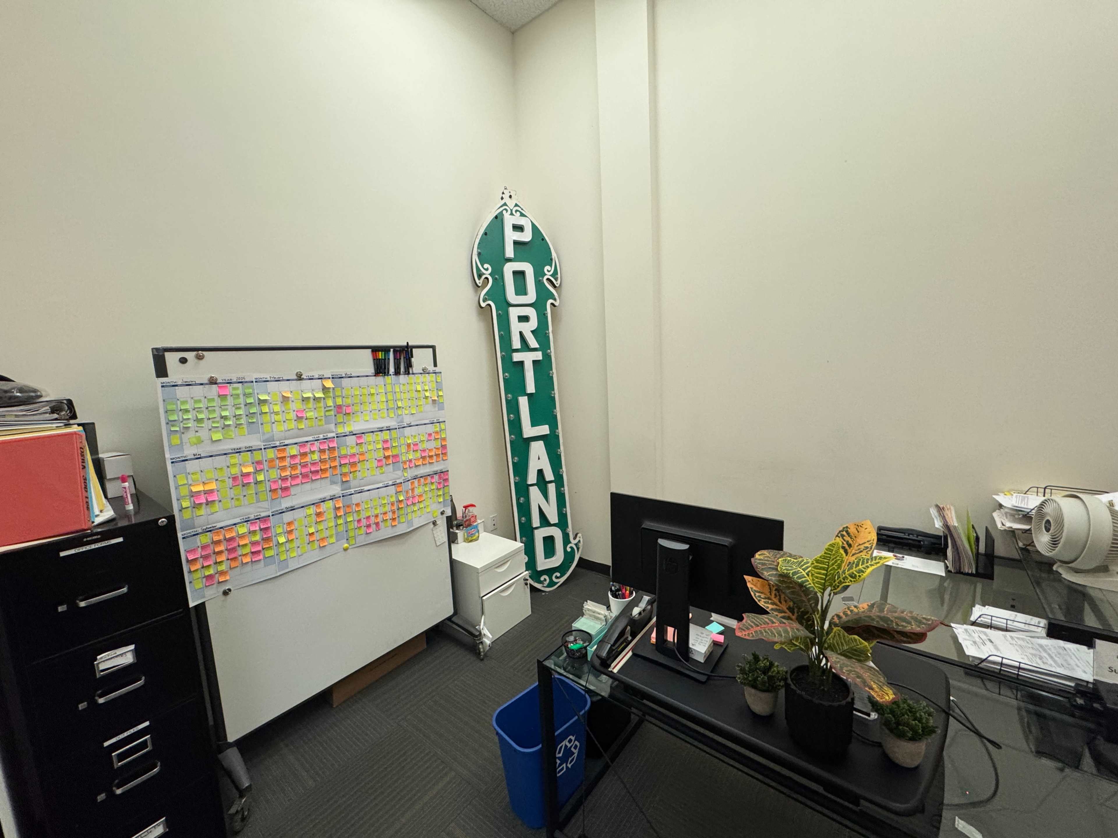 The image shows an office space with a large "PORTLAND" sign on the wall, a desk with a computer, a filing cabinet, and a whiteboard covered in colorful sticky notes.