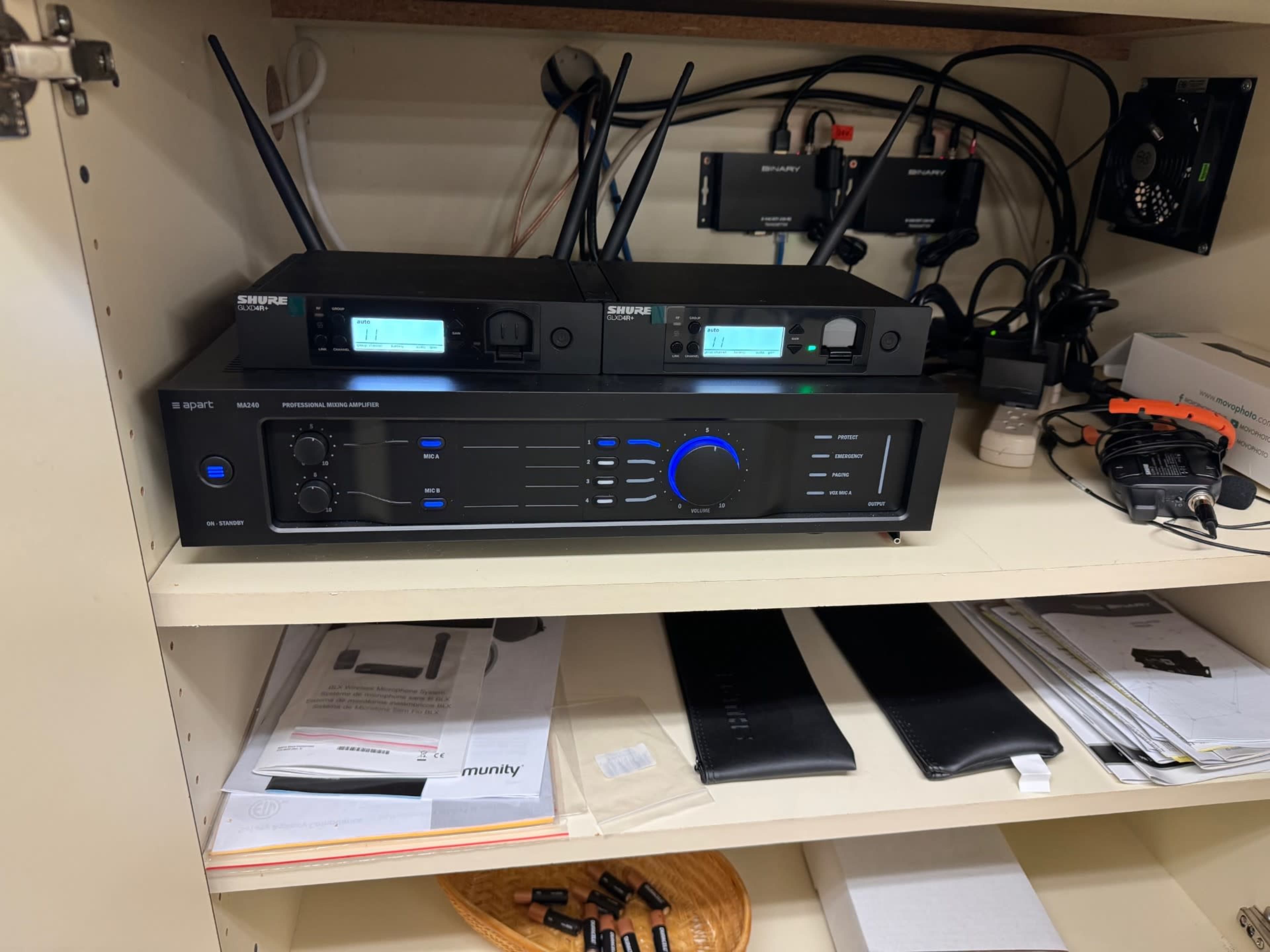 A shelf with audio equipment, including a professional audio amplifier and wireless receivers, along with various papers and accessories.