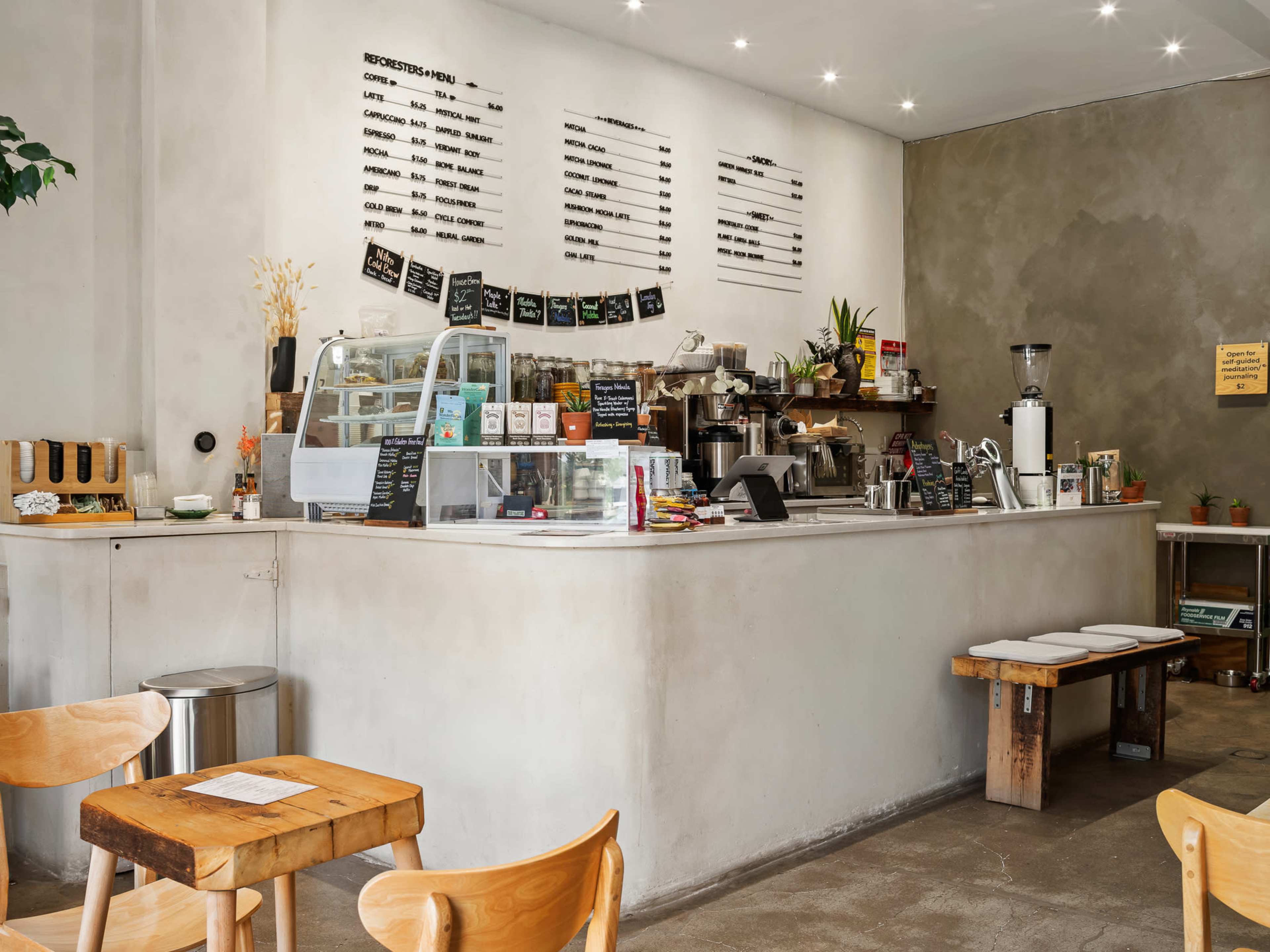The image shows a modern café interior with a minimalist design, featuring a service counter filled with baked goods and drinks, surrounded by wooden tables and chairs.
