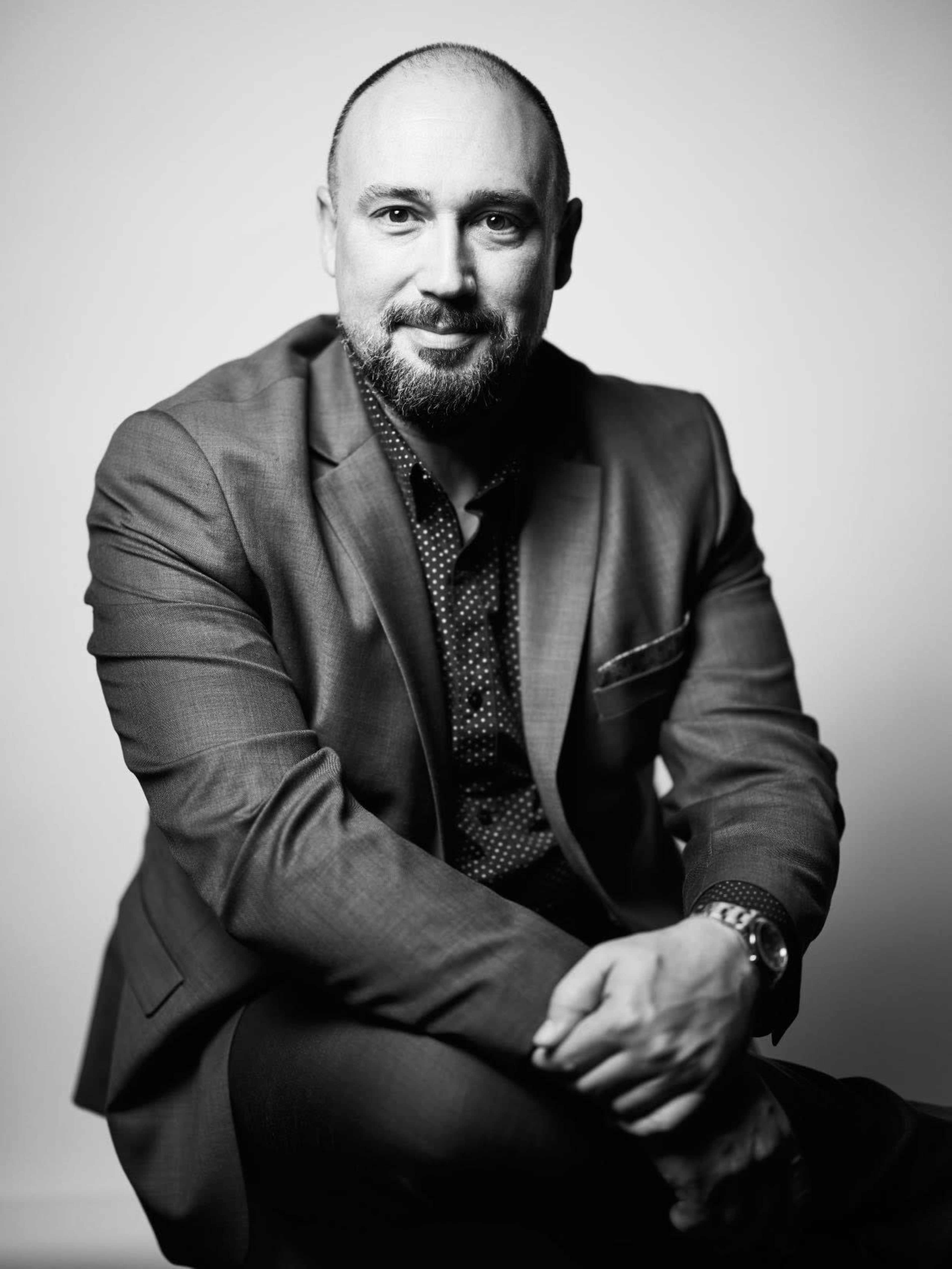 A man in a suit is seated with one arm resting on his knee and a serious expression.