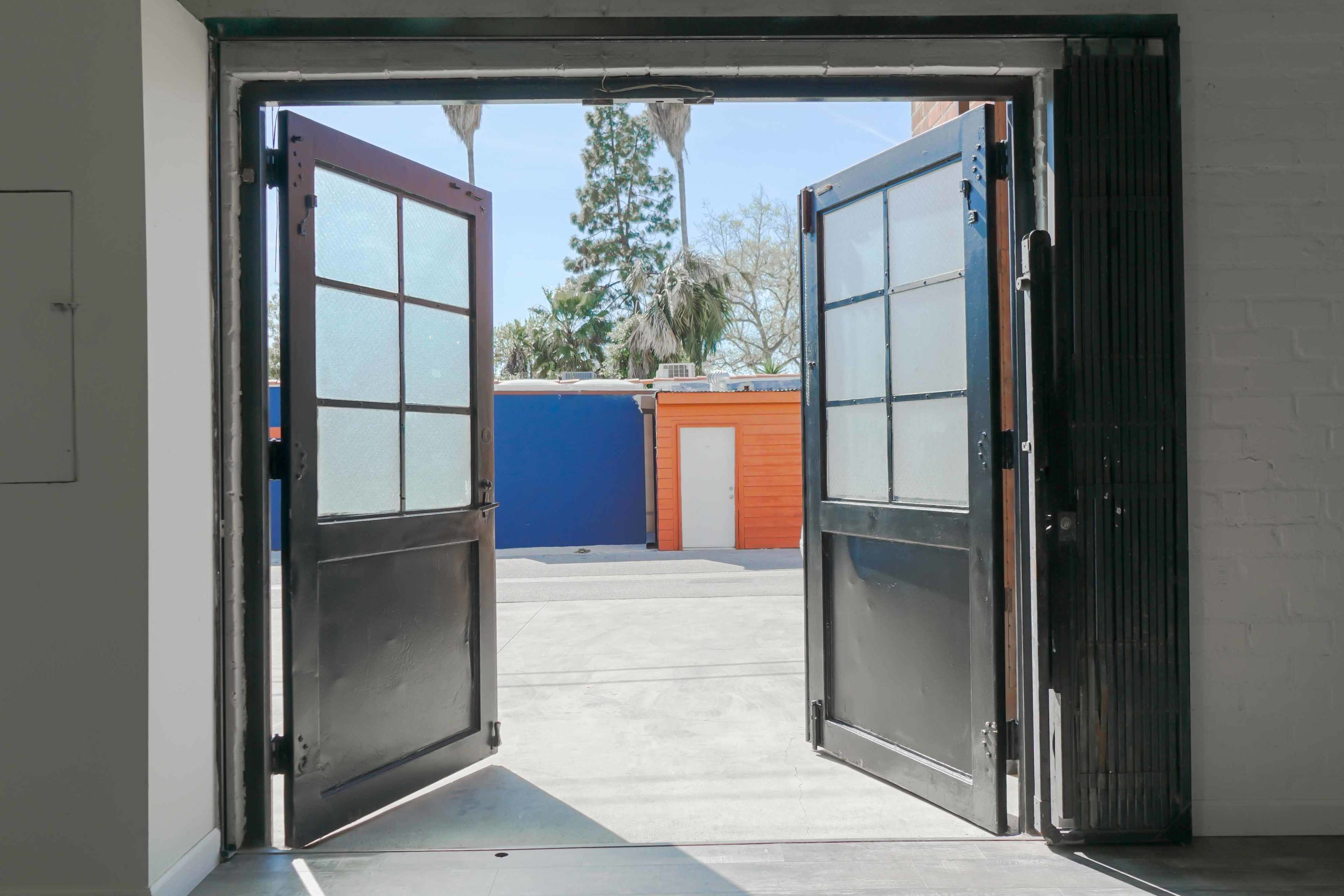 The image shows a pair of open double doors leading outside to a brightly colored courtyard with various structures.