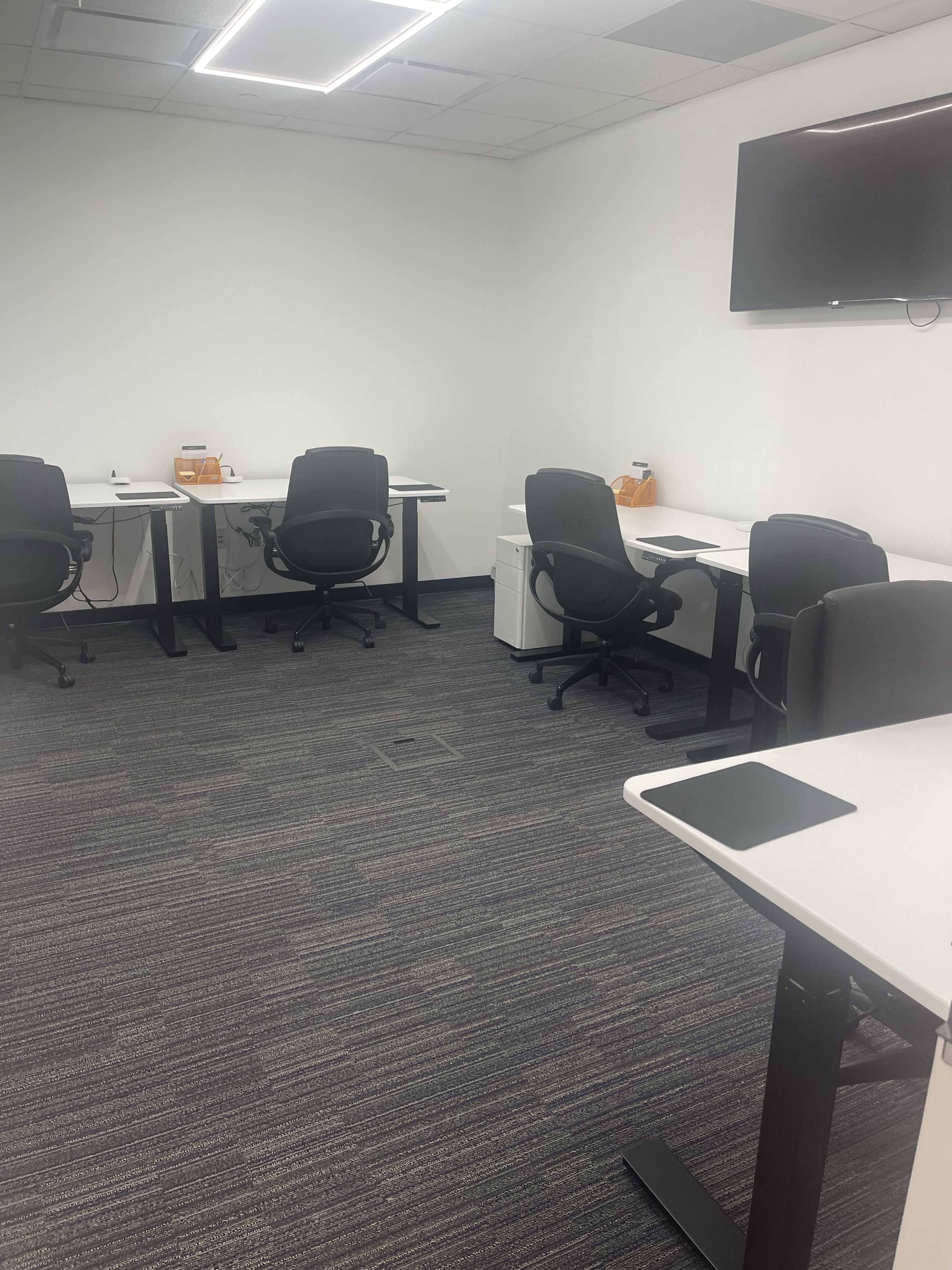 A sparsely furnished office space features several black office chairs around white desks, with a wall-mounted television and a light-colored floor carpet.