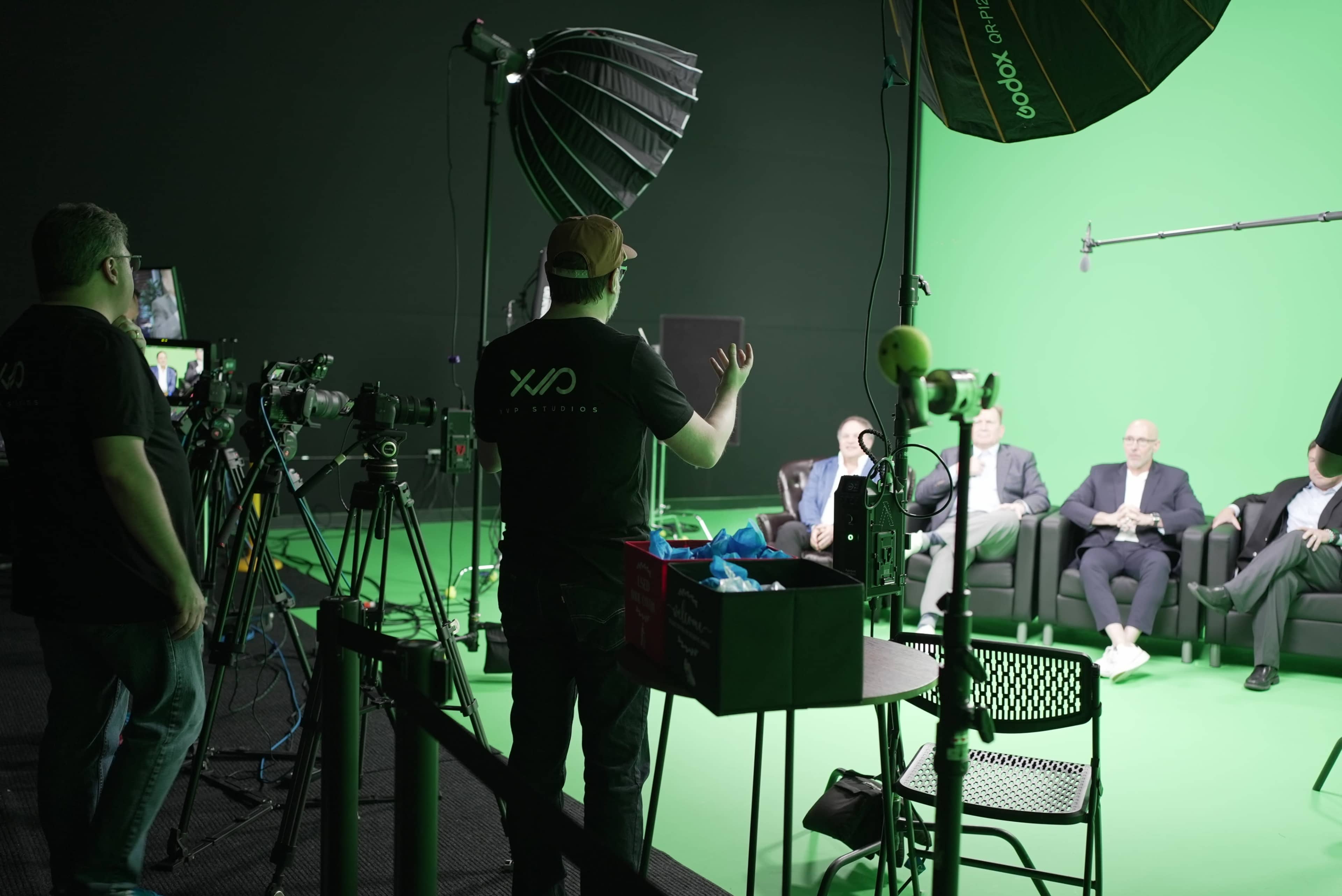 A production team sets up equipment in a green screen studio where individuals are seated on a couch.