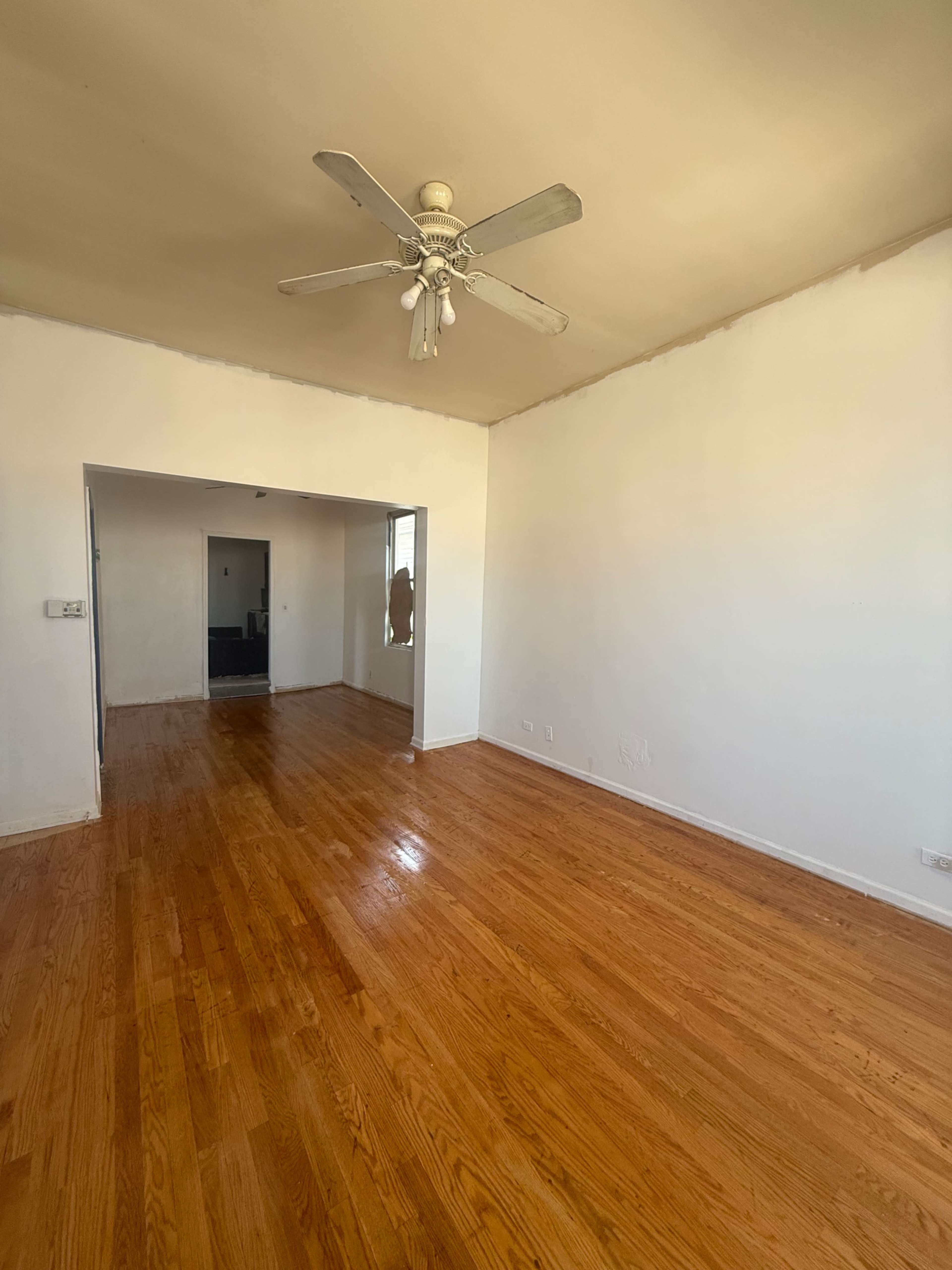 The image shows an empty room with hardwood floors, a ceiling fan, and white walls, leading to an adjoining hallway.