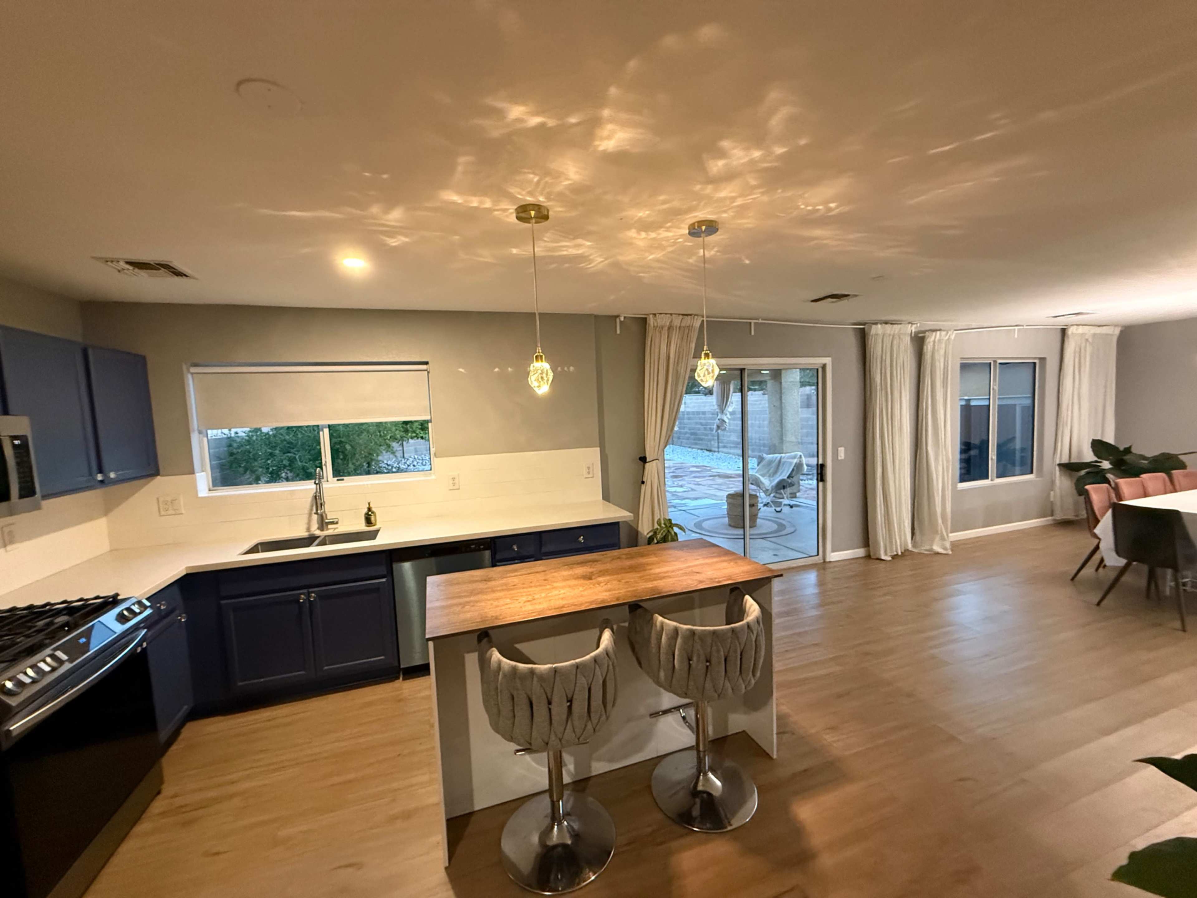 The image shows a modern kitchen with blue cabinets, a central wooden island with barstools, and a large window overlooking a patio area.