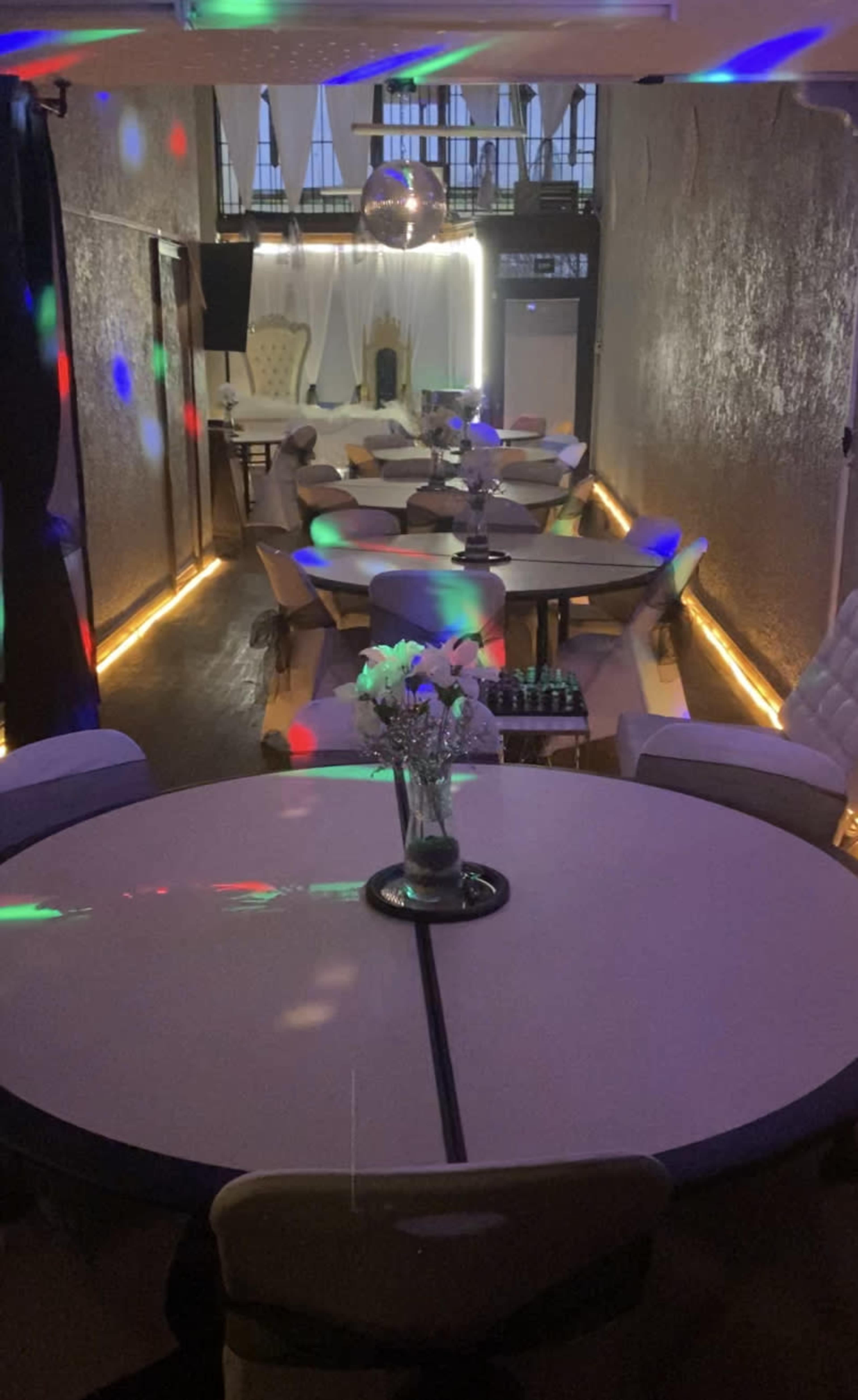 A dimly lit venue with round tables arranged in rows, decorated with floral centerpieces and illuminated by colorful lights.