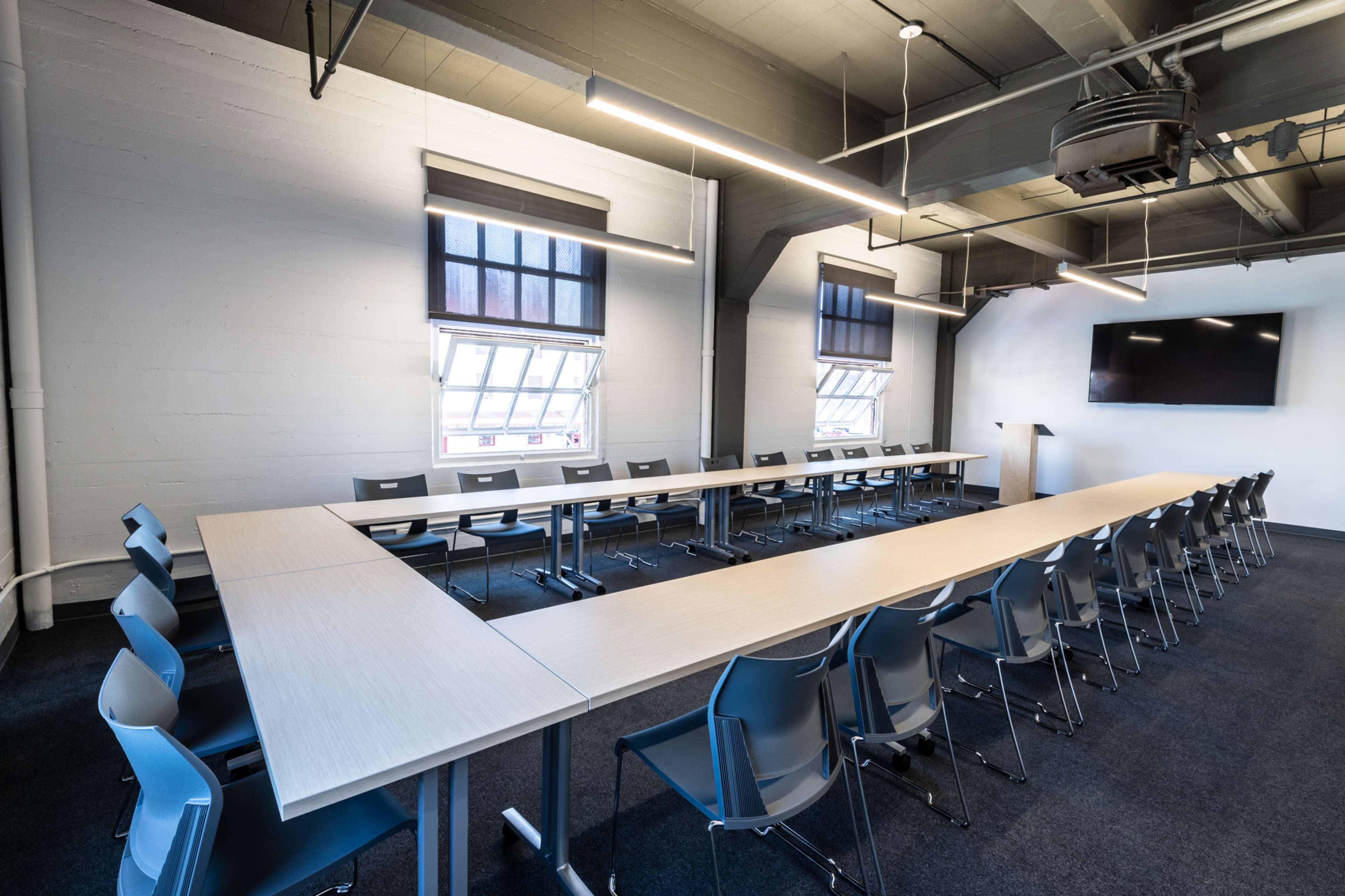 A large conference room features a long rectangular table surrounded by chairs, with windows and a screen on the walls.