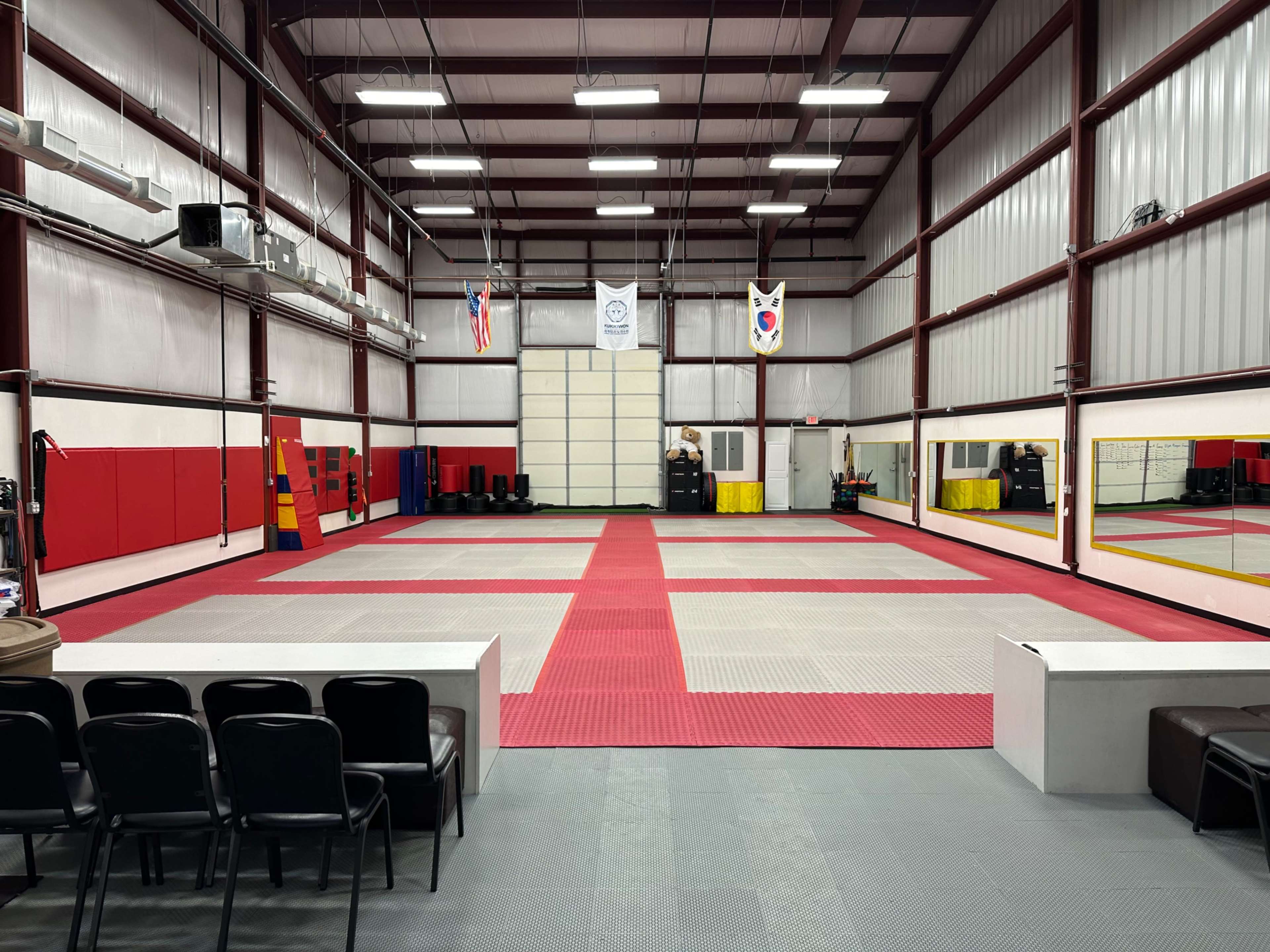 A spacious martial arts training facility with red and gray mats, mirrors along one wall, and padded equipment placed in the background.