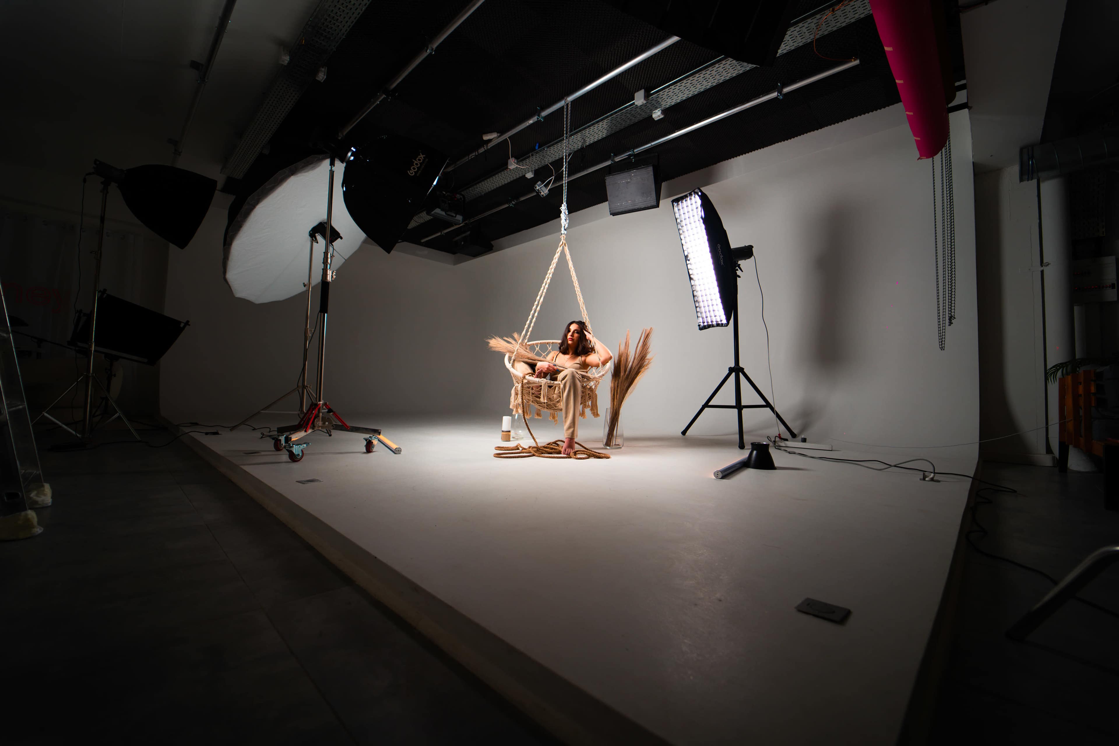 A person sits on a swing suspended from the ceiling in a well-lit studio with various lighting equipment.