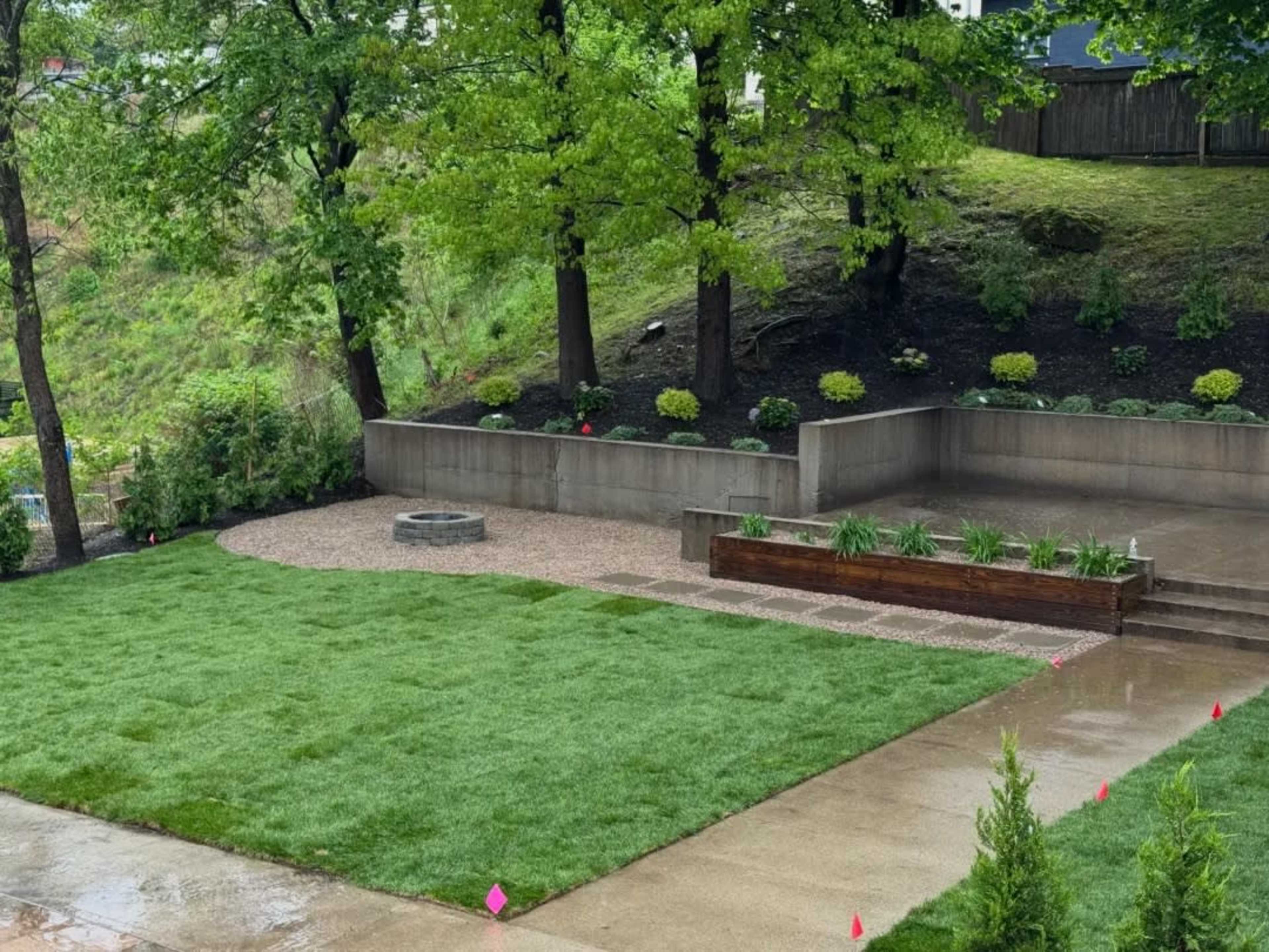 The image shows a landscaped outdoor area featuring a grassy lawn, a gravel seating area with a fire pit, and raised planters surrounded by trees.