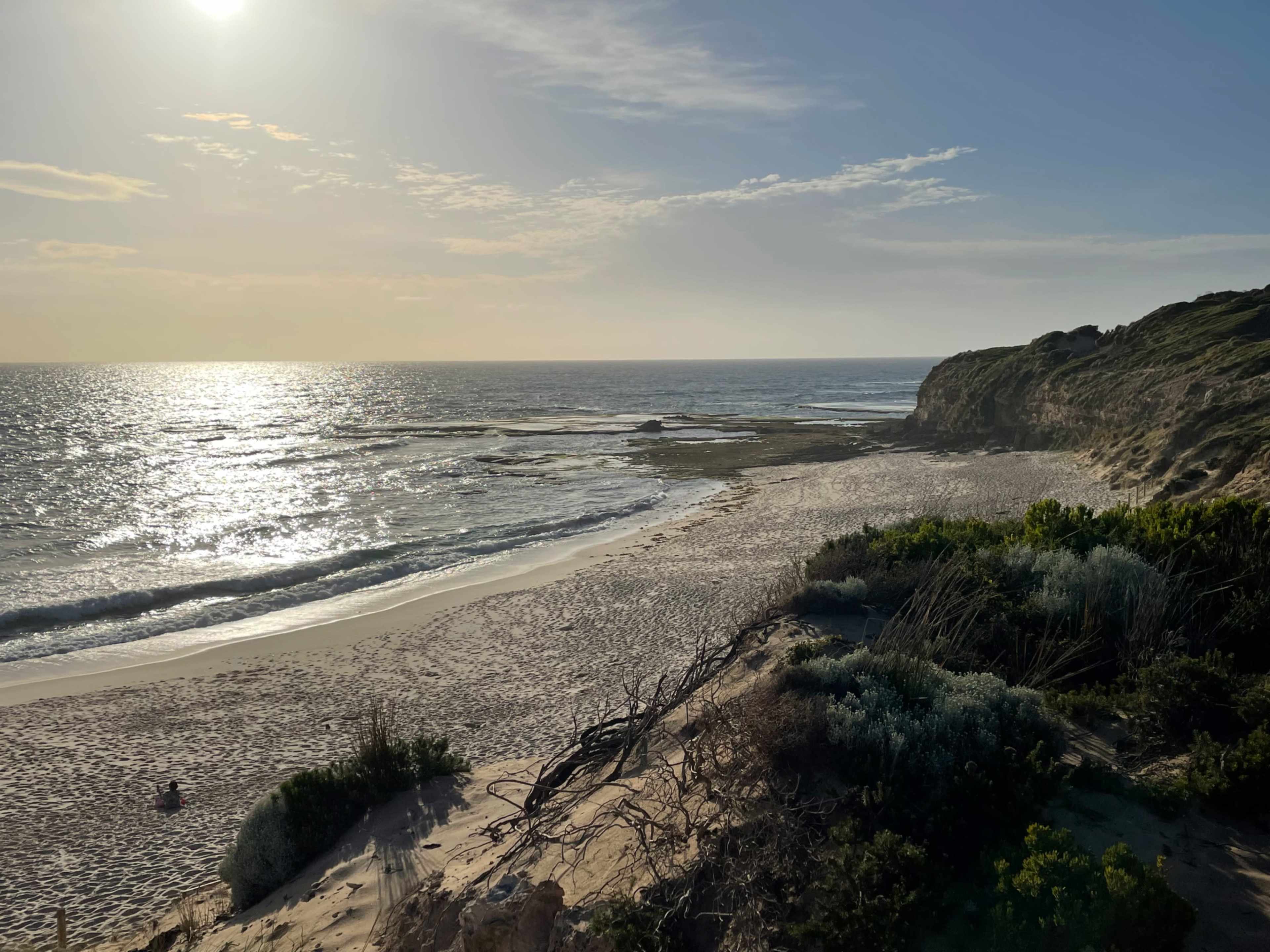 A coastal landscape with gentle waves lapping at a sandy beach, surrounded by low vegetation and cliffs under a sunlit sky.