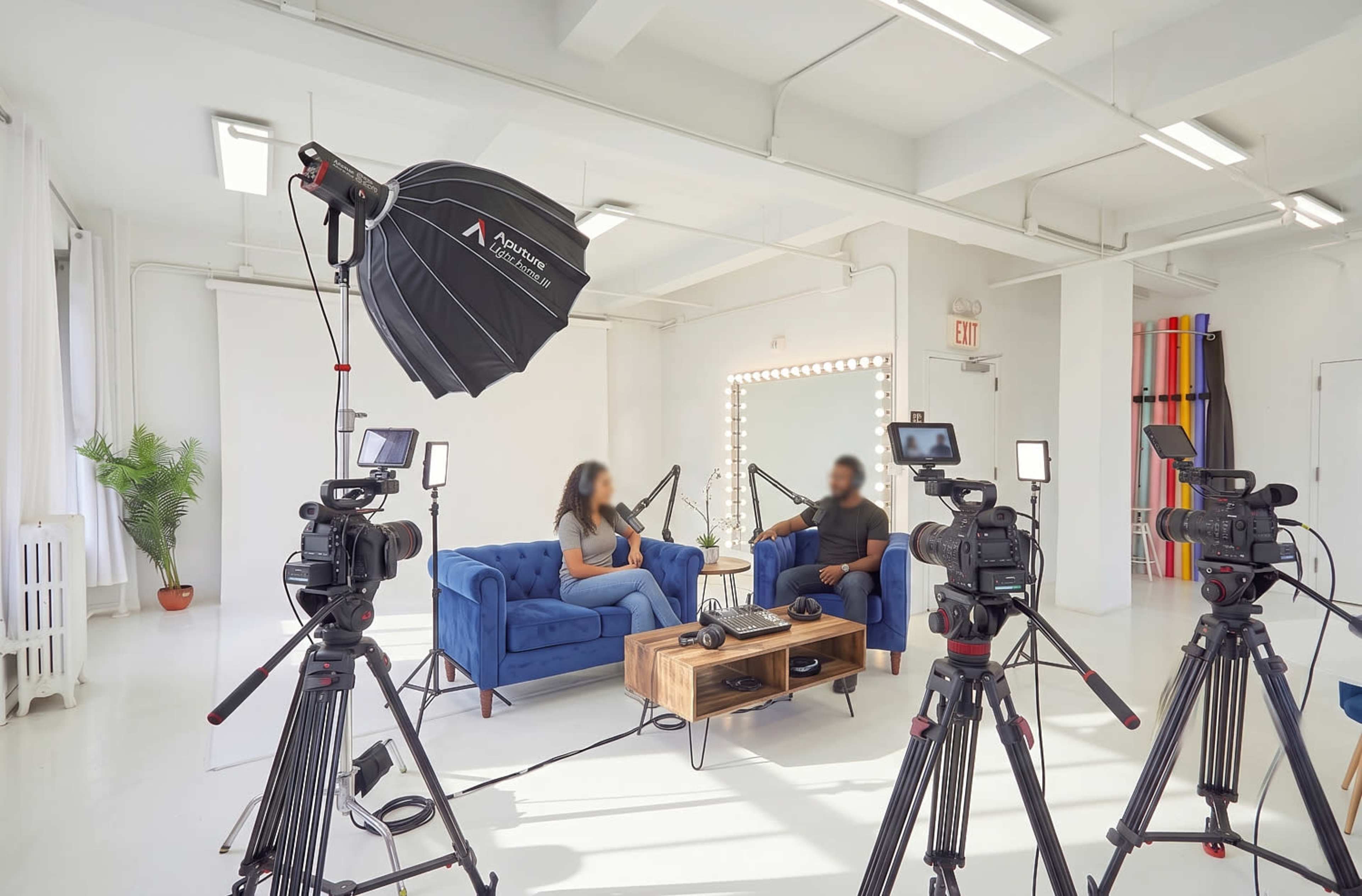 The image shows a modern studio setup for an interview, featuring two people seated on blue couches, surrounded by multiple cameras and lighting equipment.