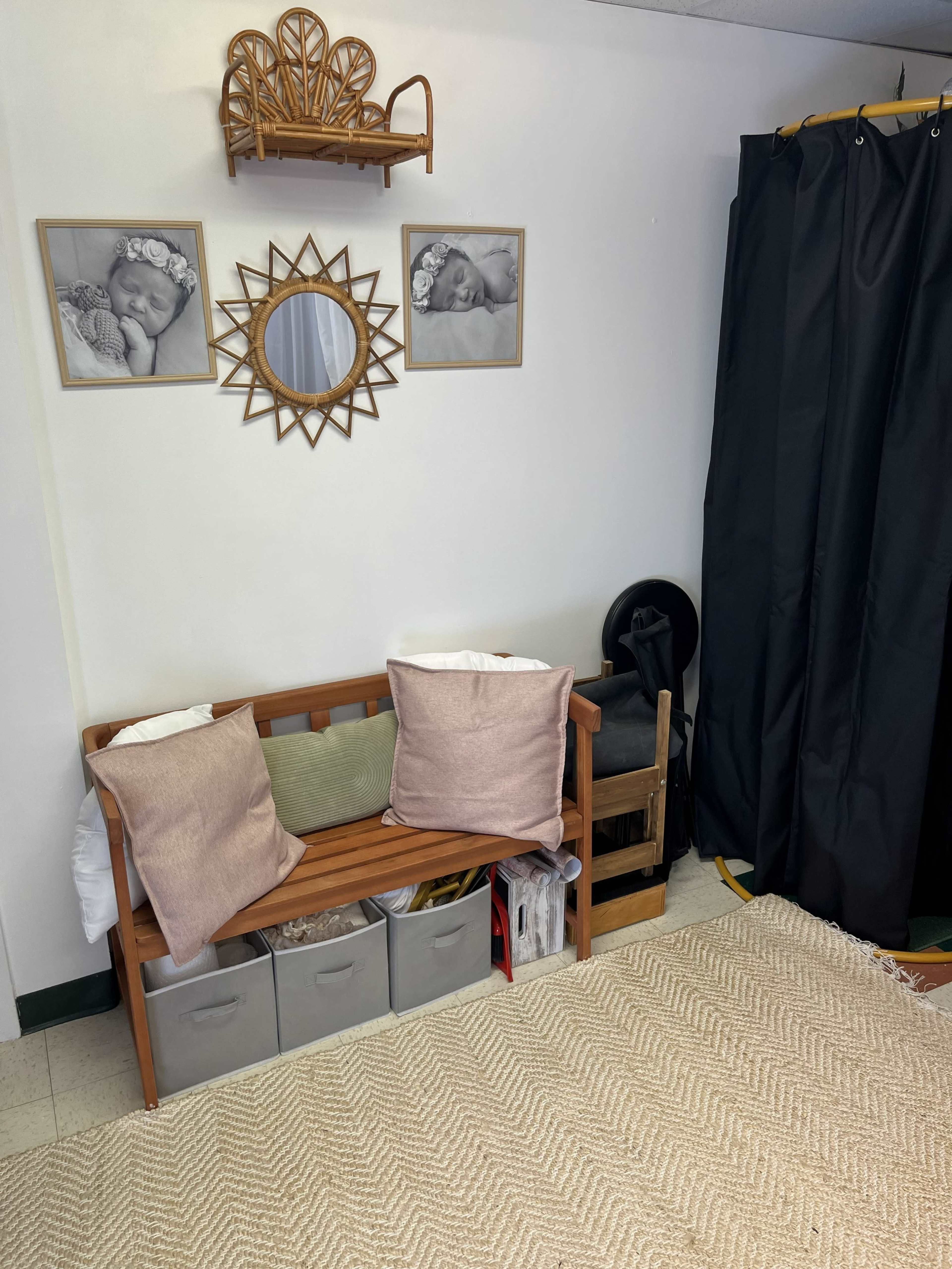 The image shows a small room featuring a wooden bench with pillows, two framed photographs on the wall, a round mirror, storage boxes, and a curtain dividing the space.