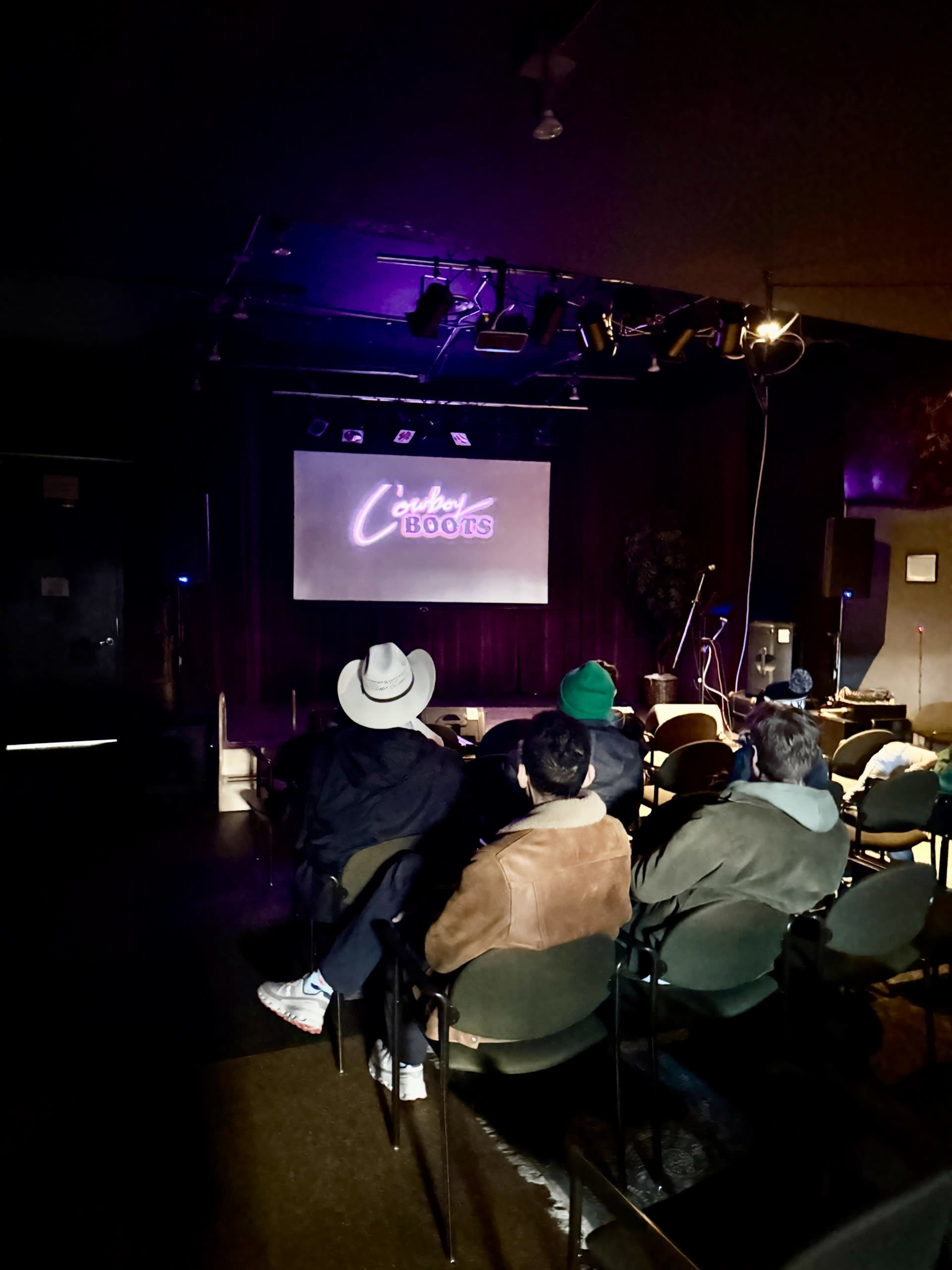 A small audience sits in a dimly lit room facing a screen that reads "Cozy Books."