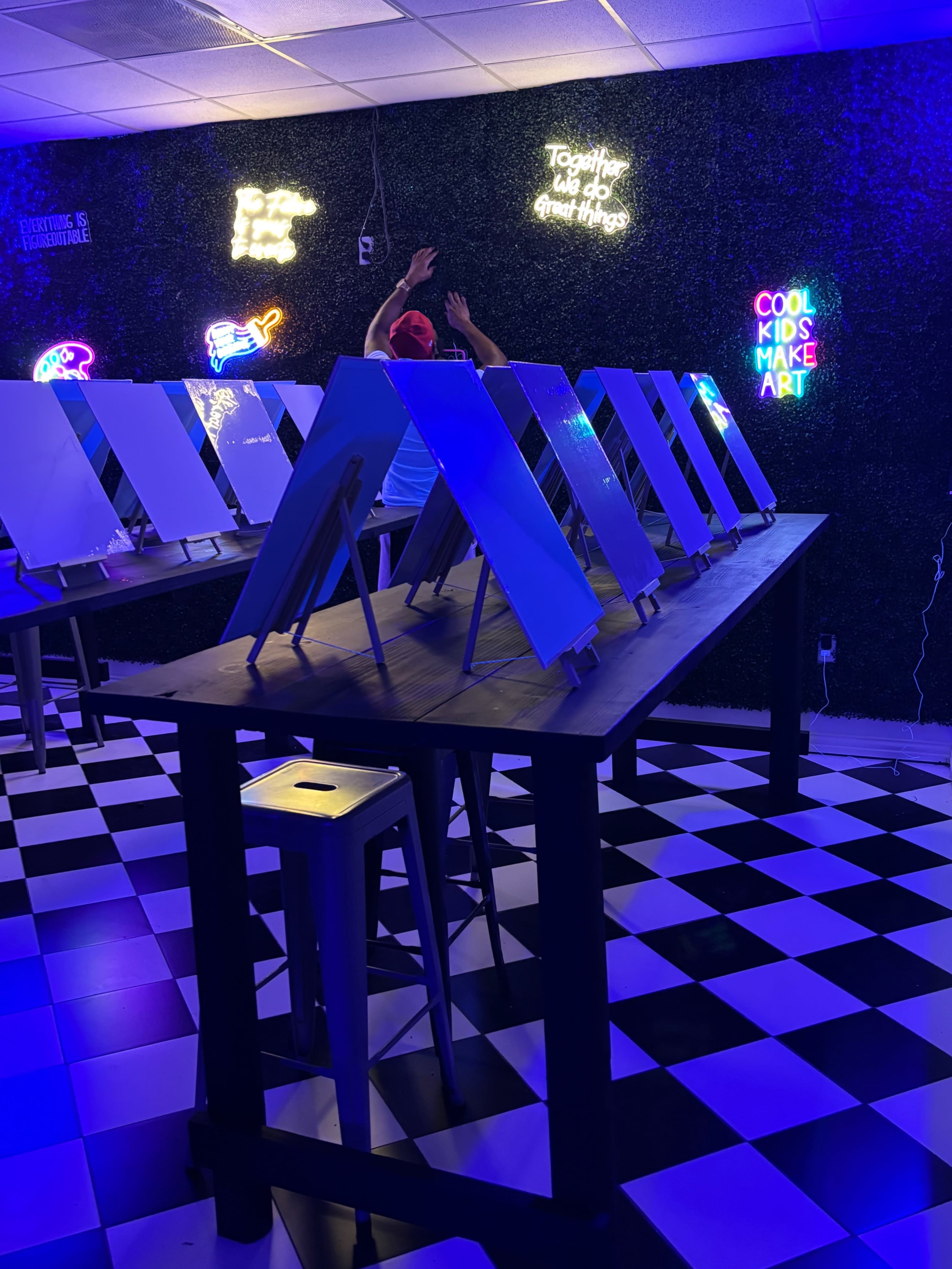 The image shows a brightly lit art studio with several easels set up on tables, surrounded by colorful neon signs.