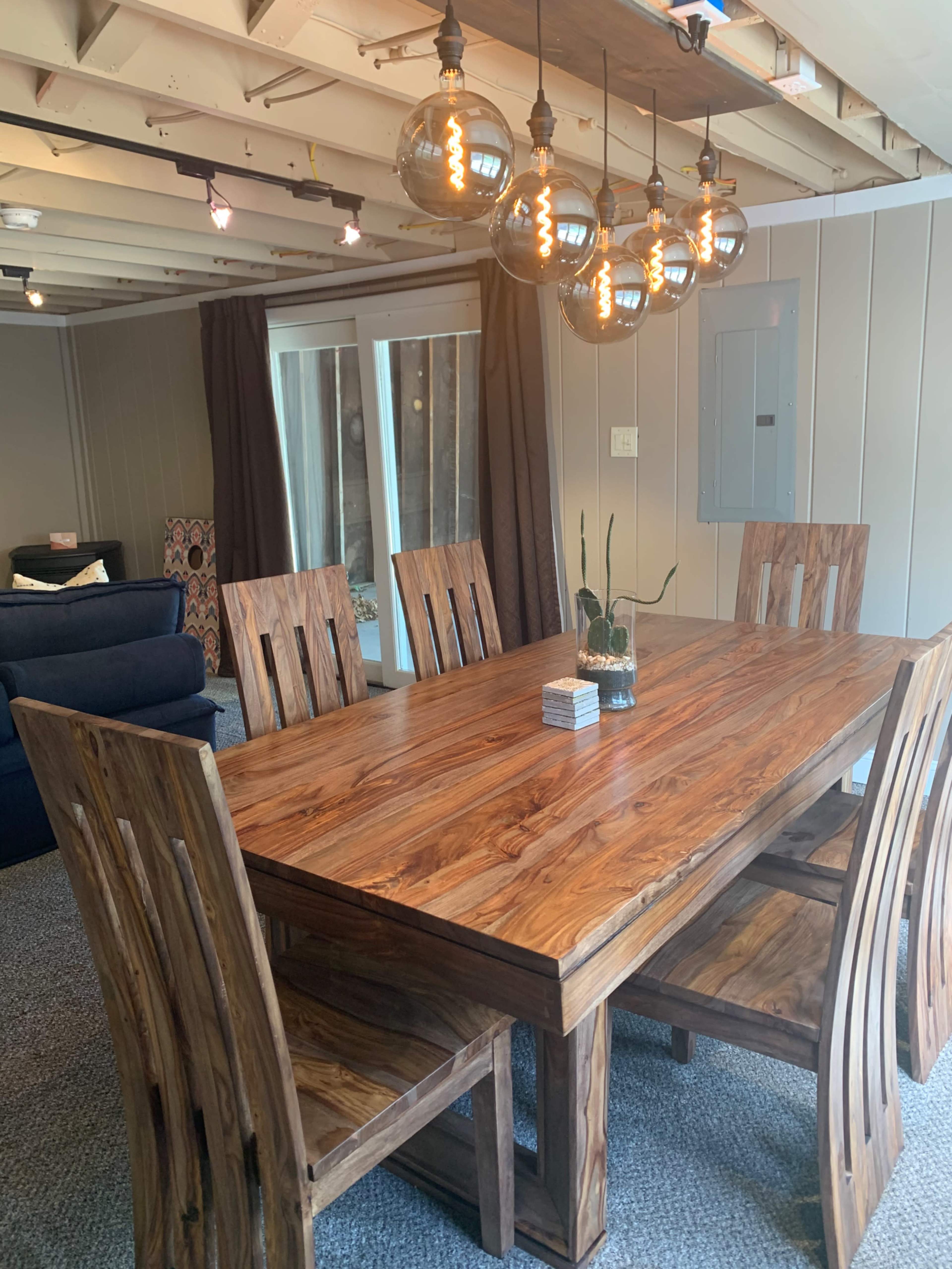 A wooden dining table surrounded by four chairs, with pendant lights hanging above in a room featuring a sofa and large windows.