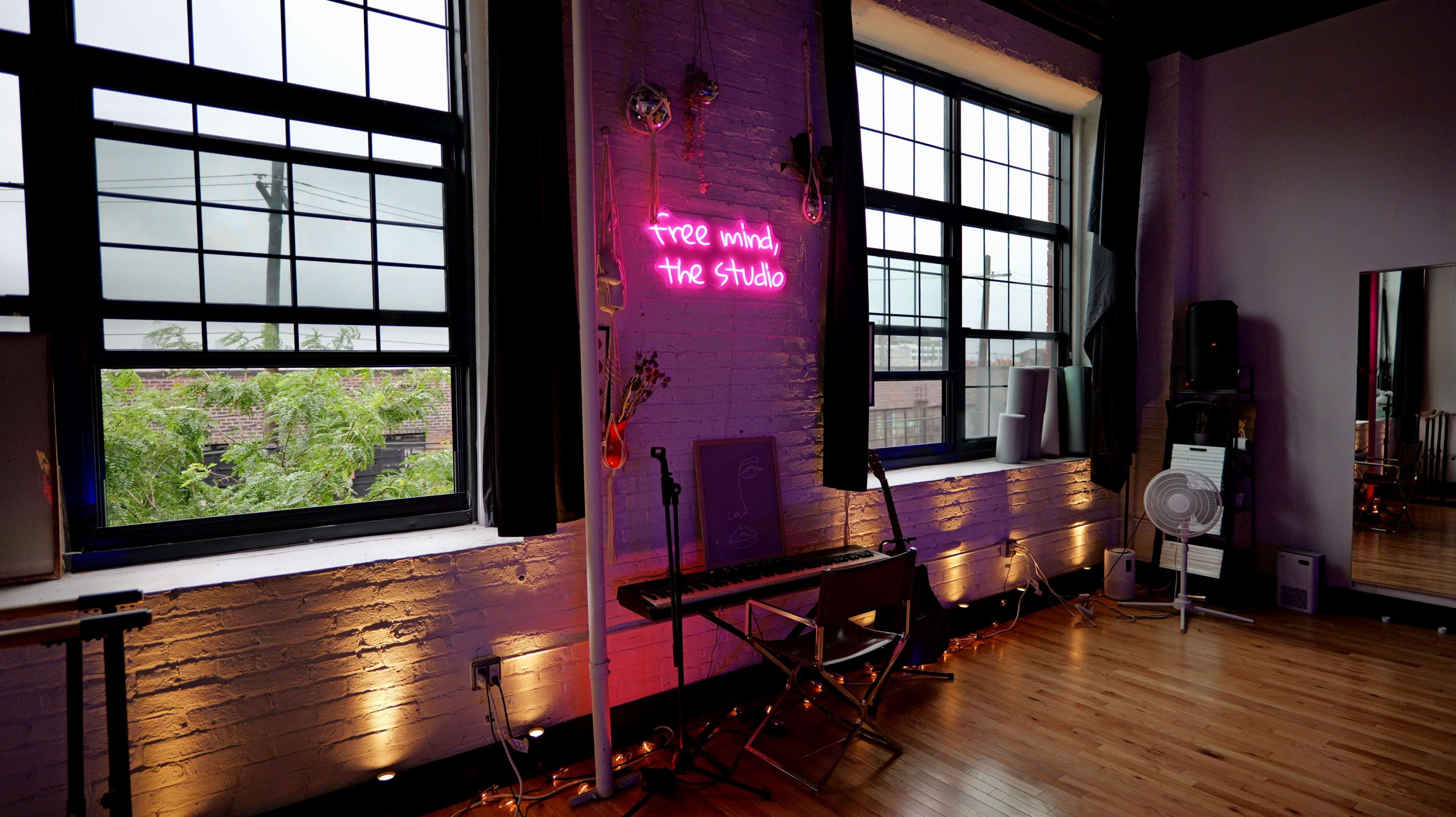 A music studio is illuminated by a neon sign that reads "free mind, the Studio," with large windows providing natural light and greenery visible outside.