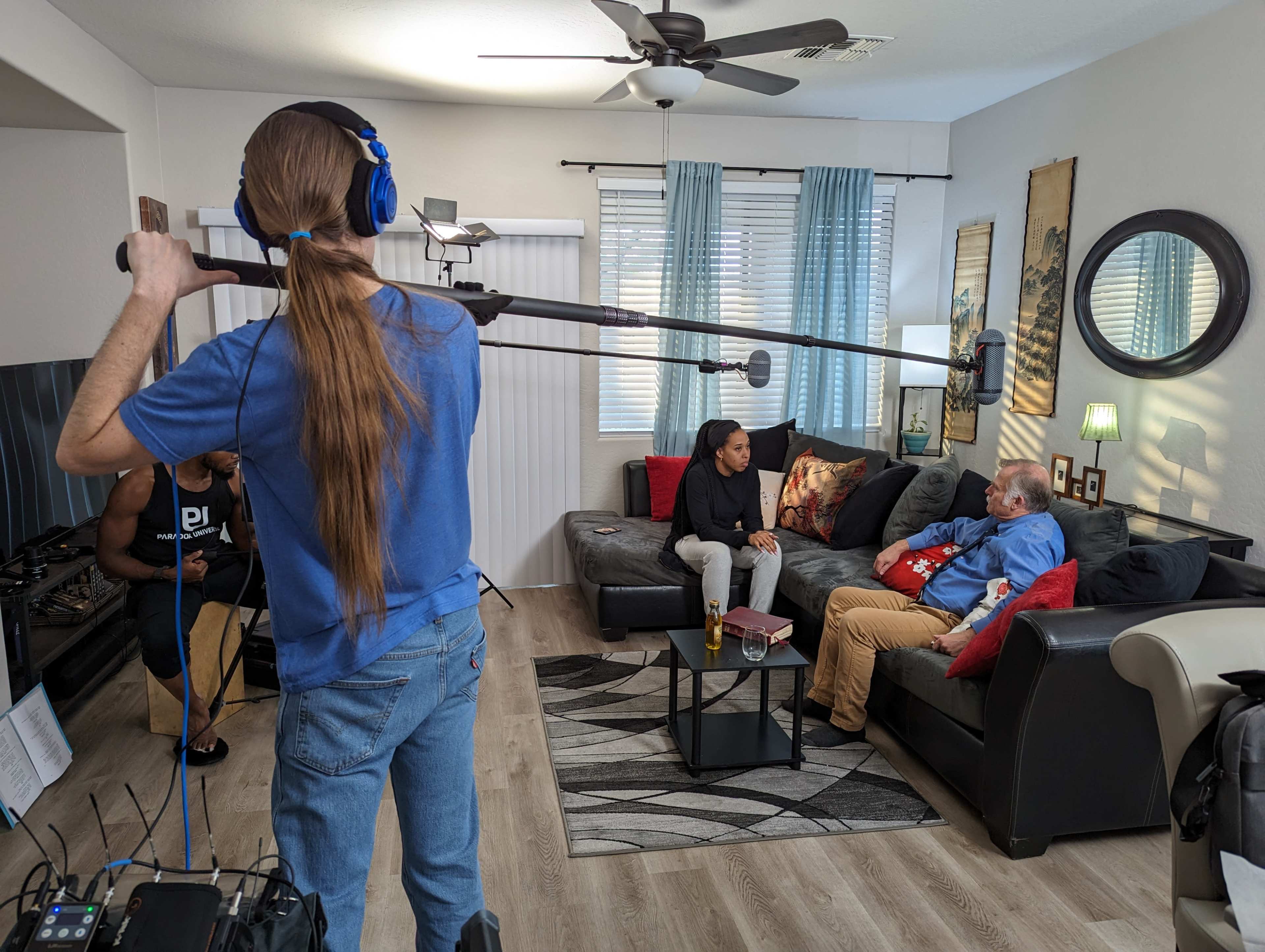 A film crew member holds a boom microphone while two actors sit on a couch in a living room set.
