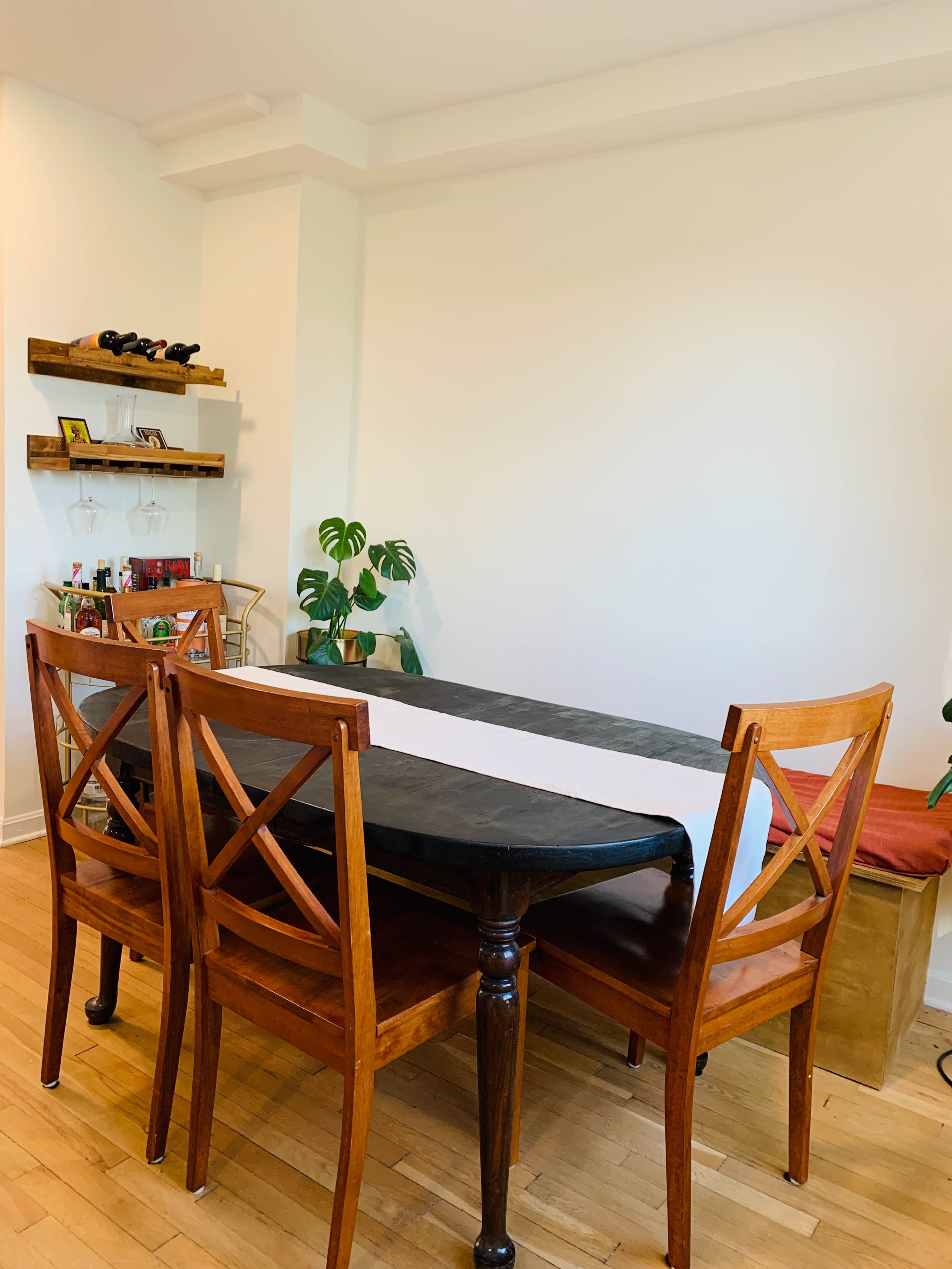 A wooden dining table with a black tabletop is surrounded by four wooden chairs in a simple dining area featuring a plant and a small shelf.