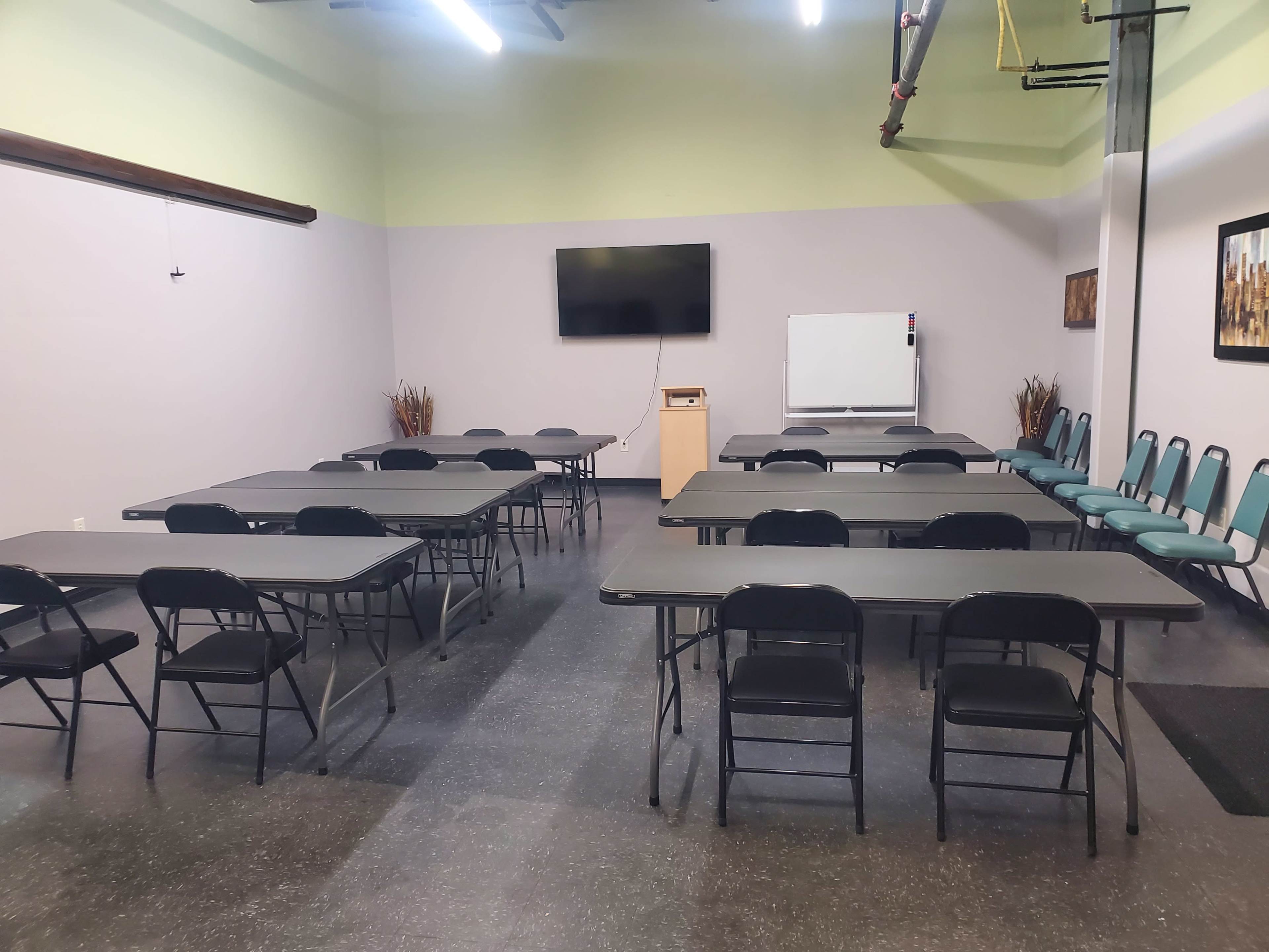 A spacious meeting room features several rows of black folding tables and chairs, with a TV and whiteboard at one end.