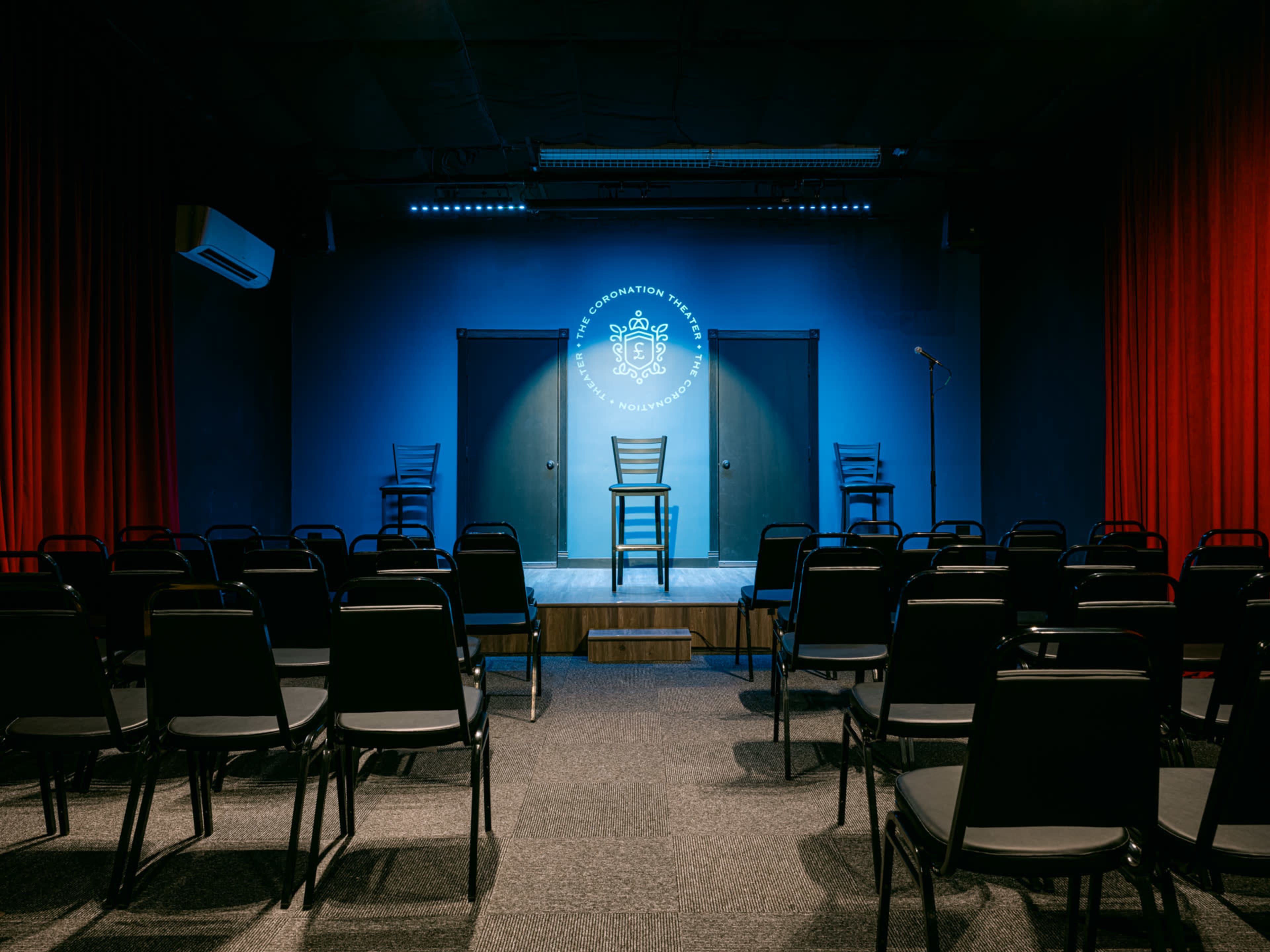 A stage with three chairs and a microphone is set up in a theater surrounded by empty black chairs facing the stage.