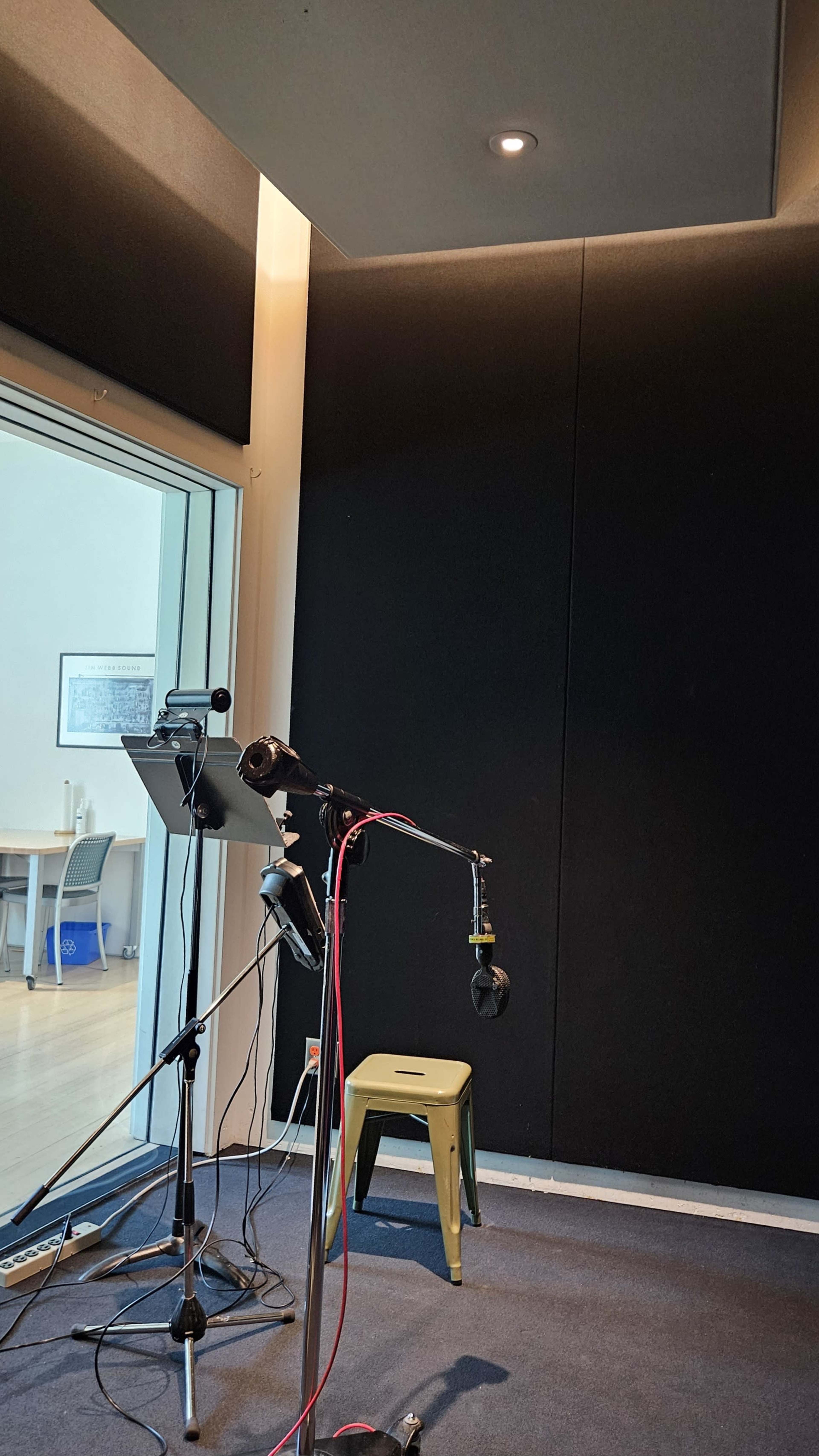 The image shows a recording studio with a microphone set up on a stand, a music stand, and a green stool in a corner, with black soundproofing walls and a glass window revealing a workspace beyond.