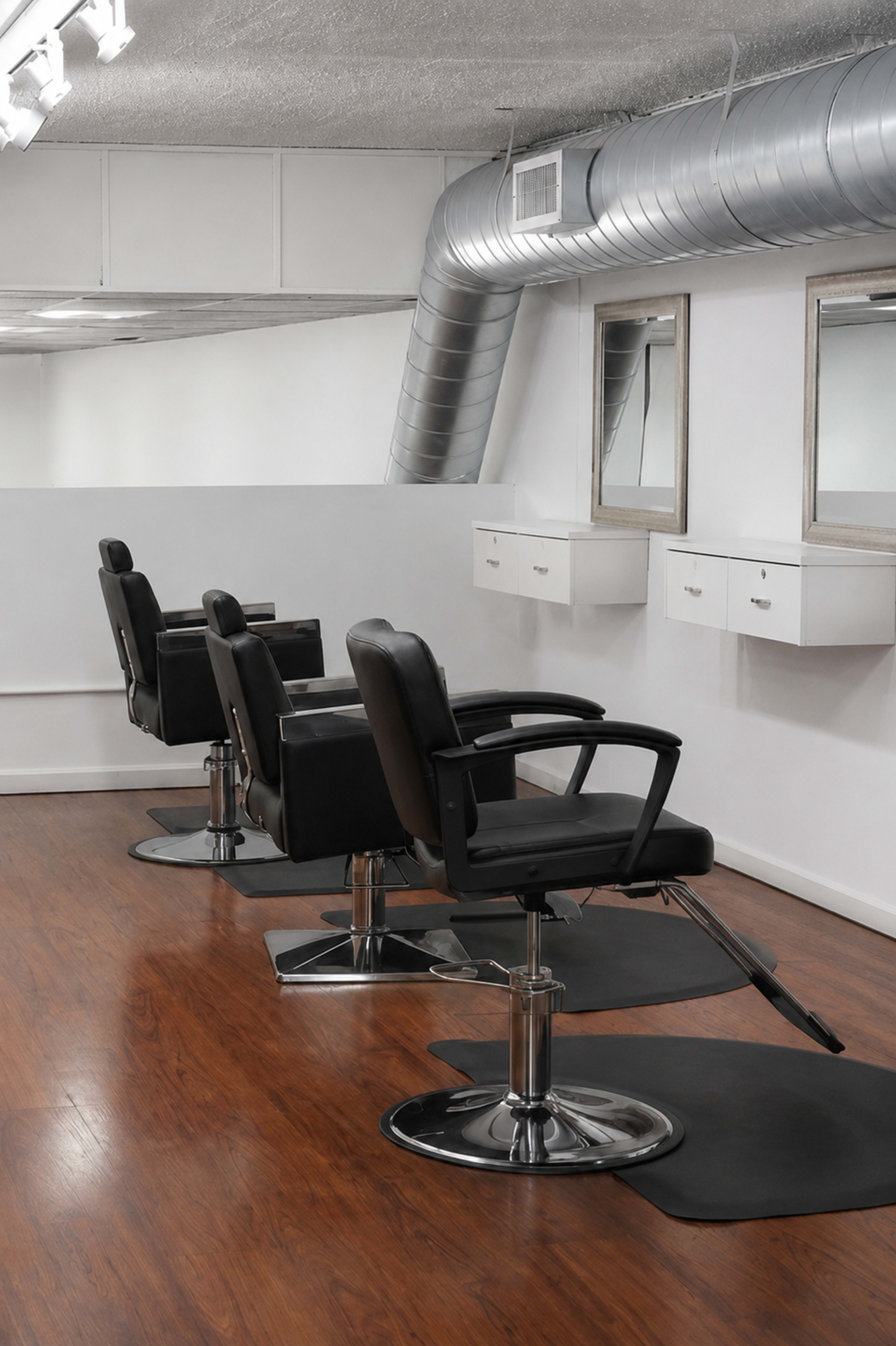 The image depicts a modern hair salon interior with three black salon chairs positioned in front of mirrors, set against a clean white wall and wooden floor.