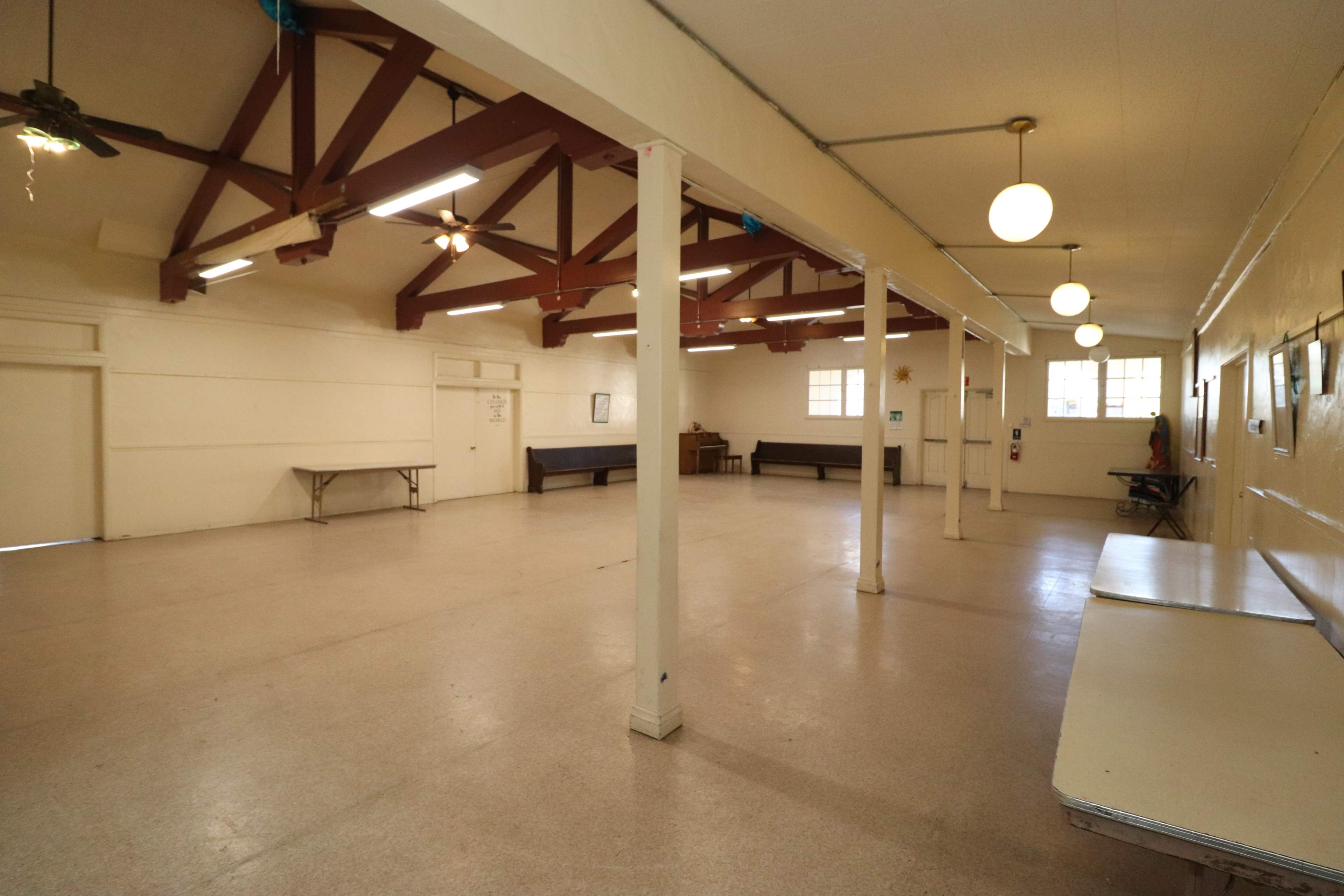 A spacious indoor room with a high ceiling, wooden beams, and several tables along the edges.