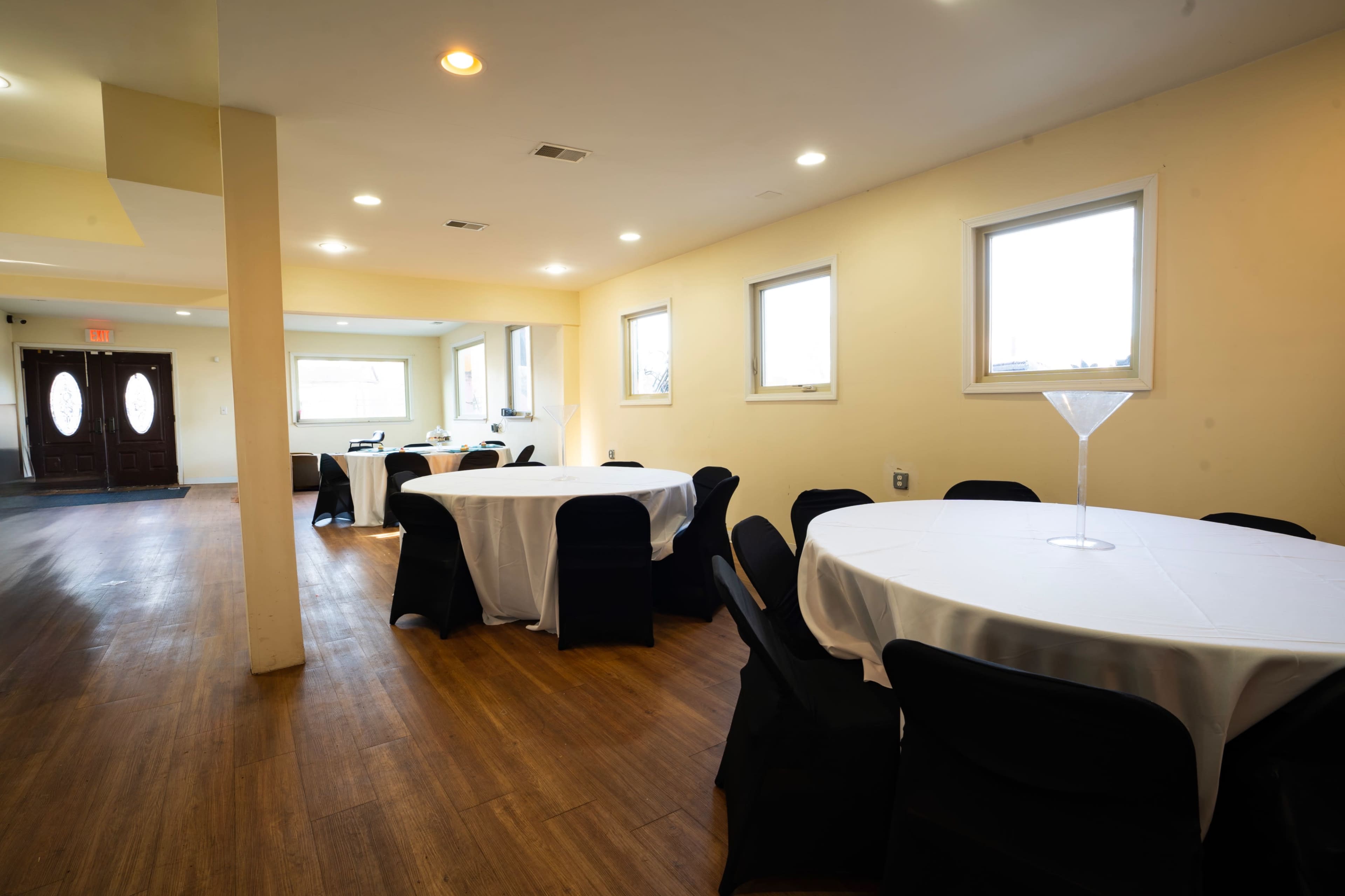 The image shows a brightly lit indoor venue featuring several round tables covered with white tablecloths and black chairs arranged along a wooden floor.