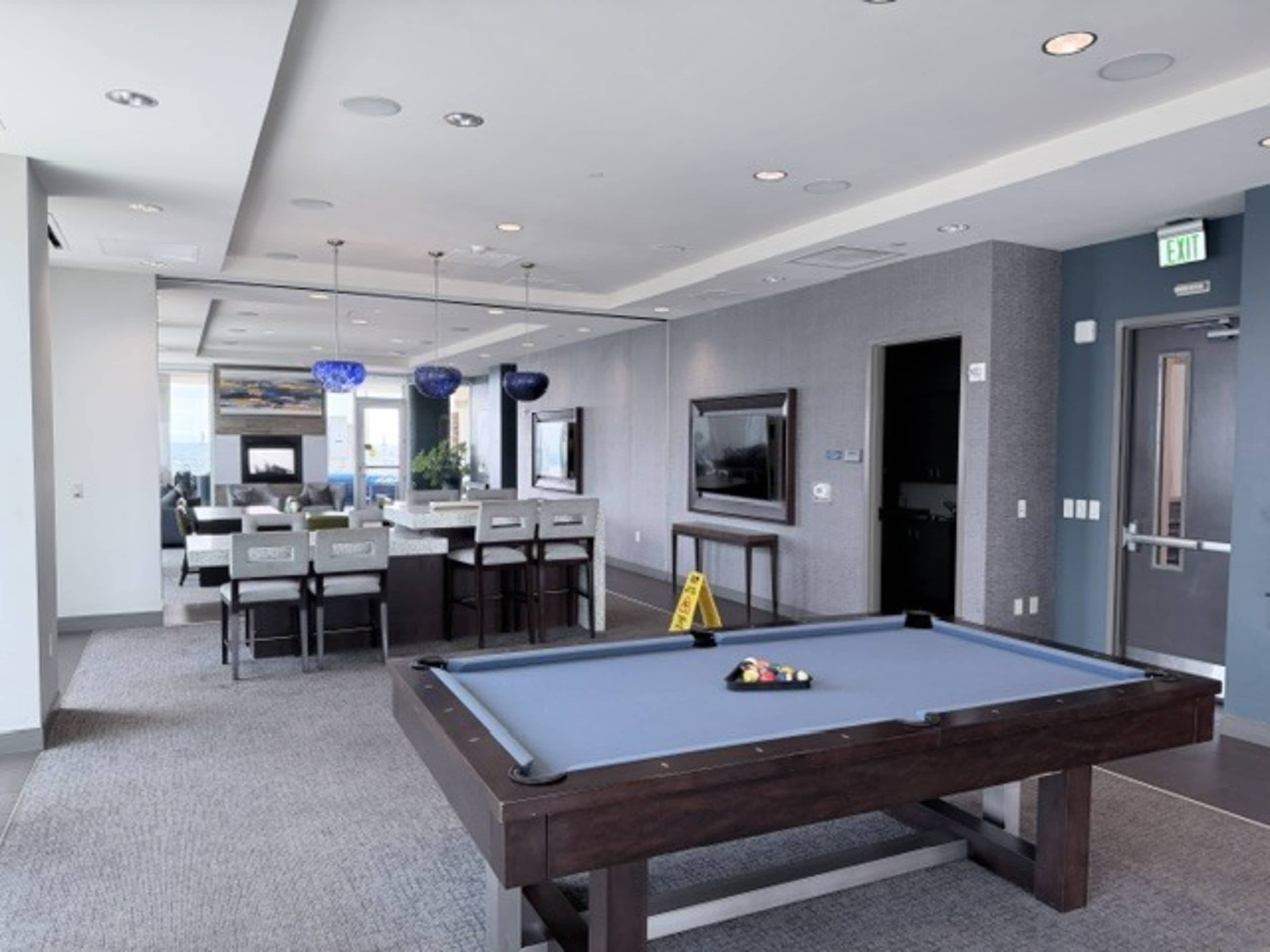 The image shows a modern recreation room featuring a billiards table in the foreground and a lounge area with seating and a television in the background.