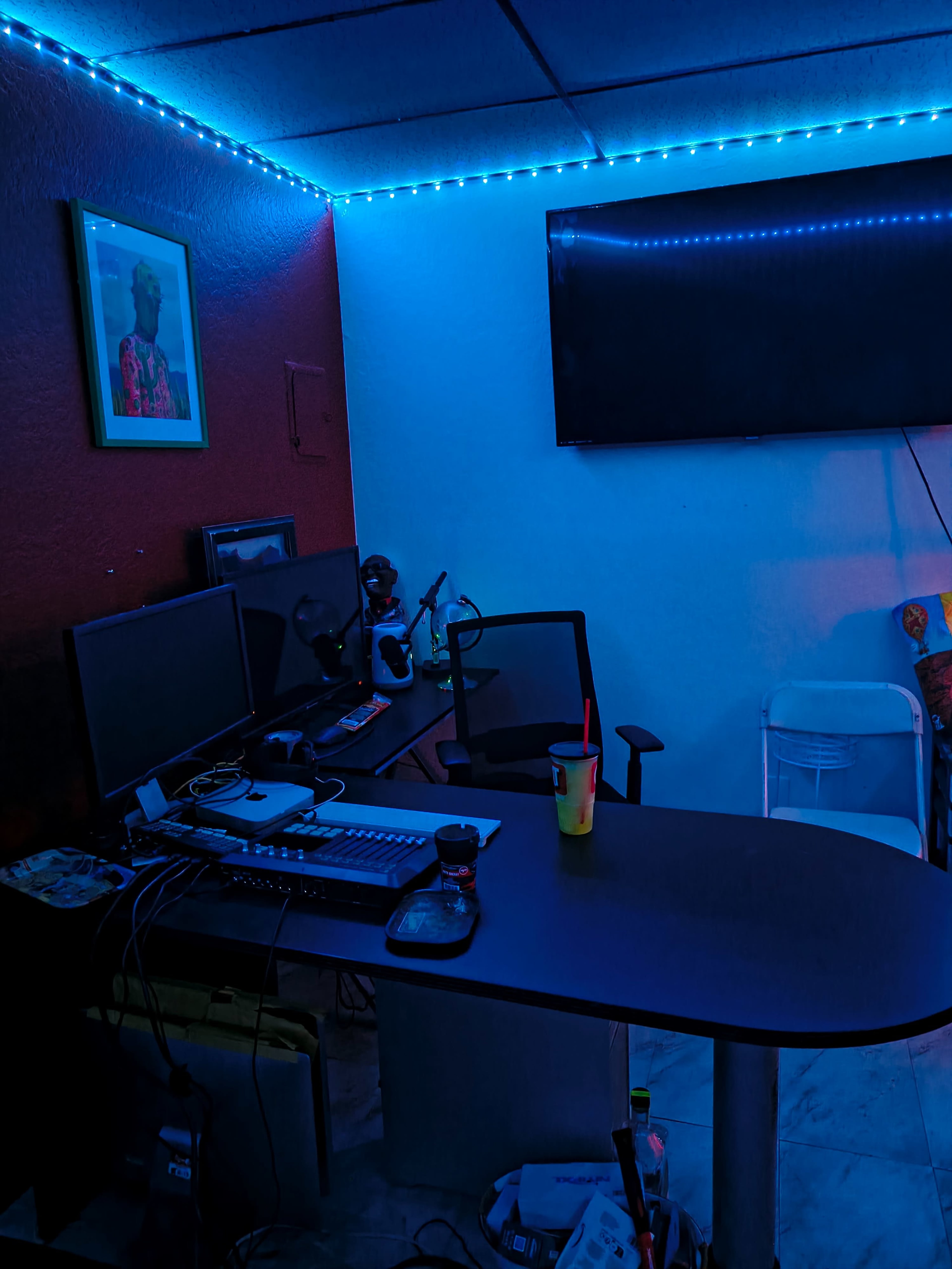 A dimly lit office space features a desk with dual monitors, a chair, and a flat-screen TV mounted on the wall, illuminated by blue LED lights.
