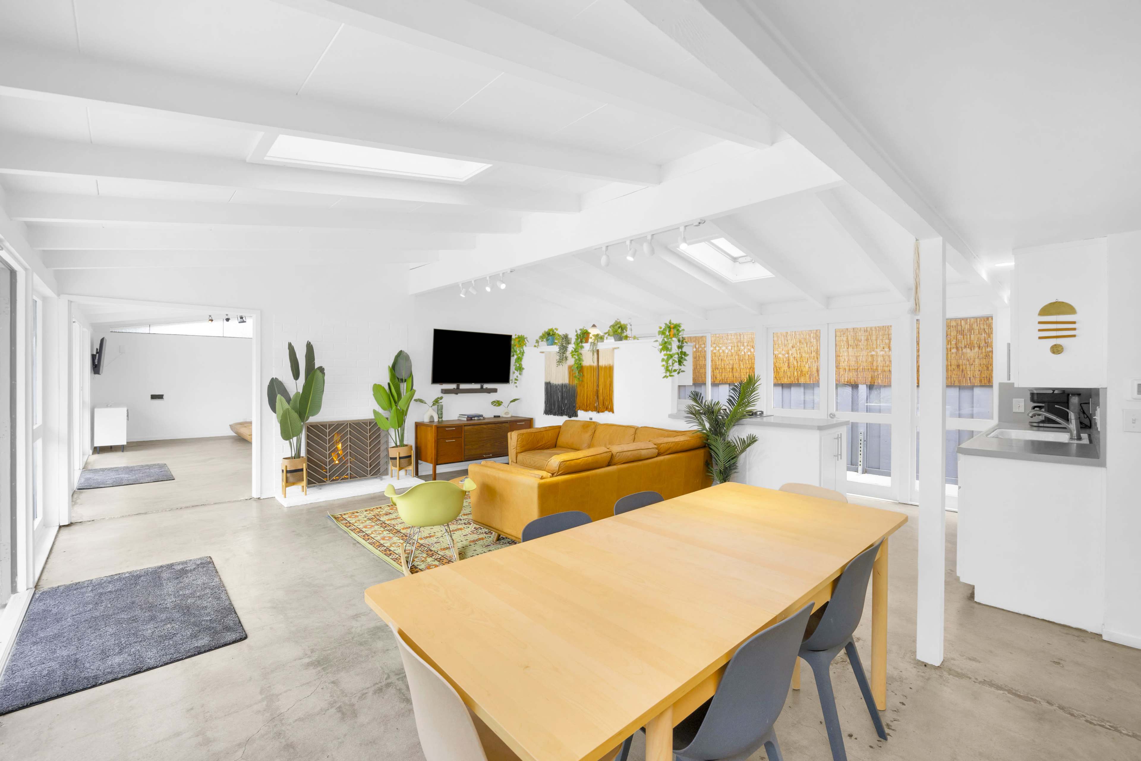 The image shows an open-concept living space with a dining area, a lounge with a patterned sofa, and large windows illuminated by natural light.