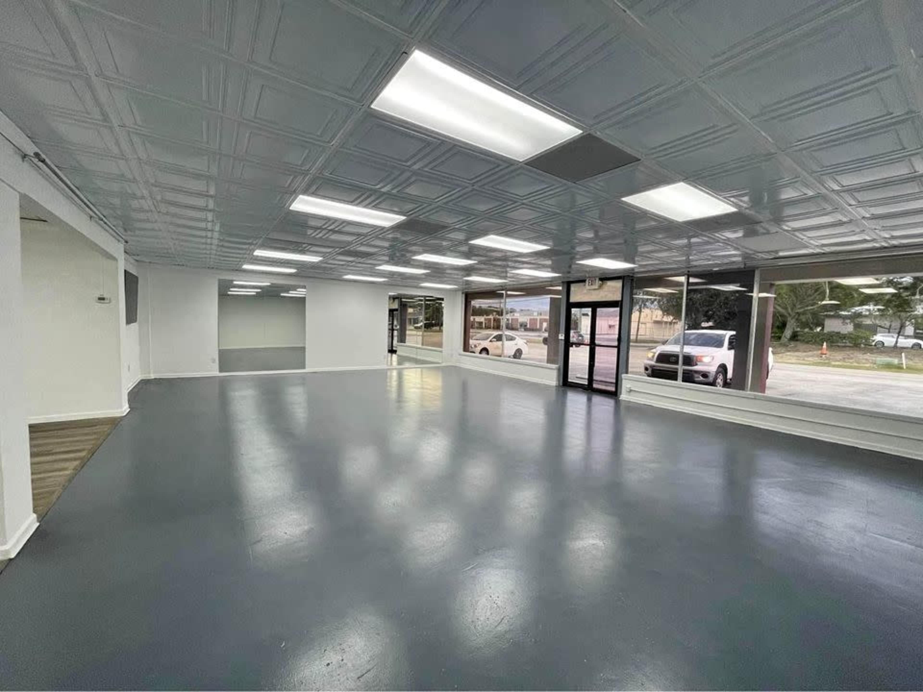 The image shows an empty commercial space with a gray floor, white walls, and large front windows.