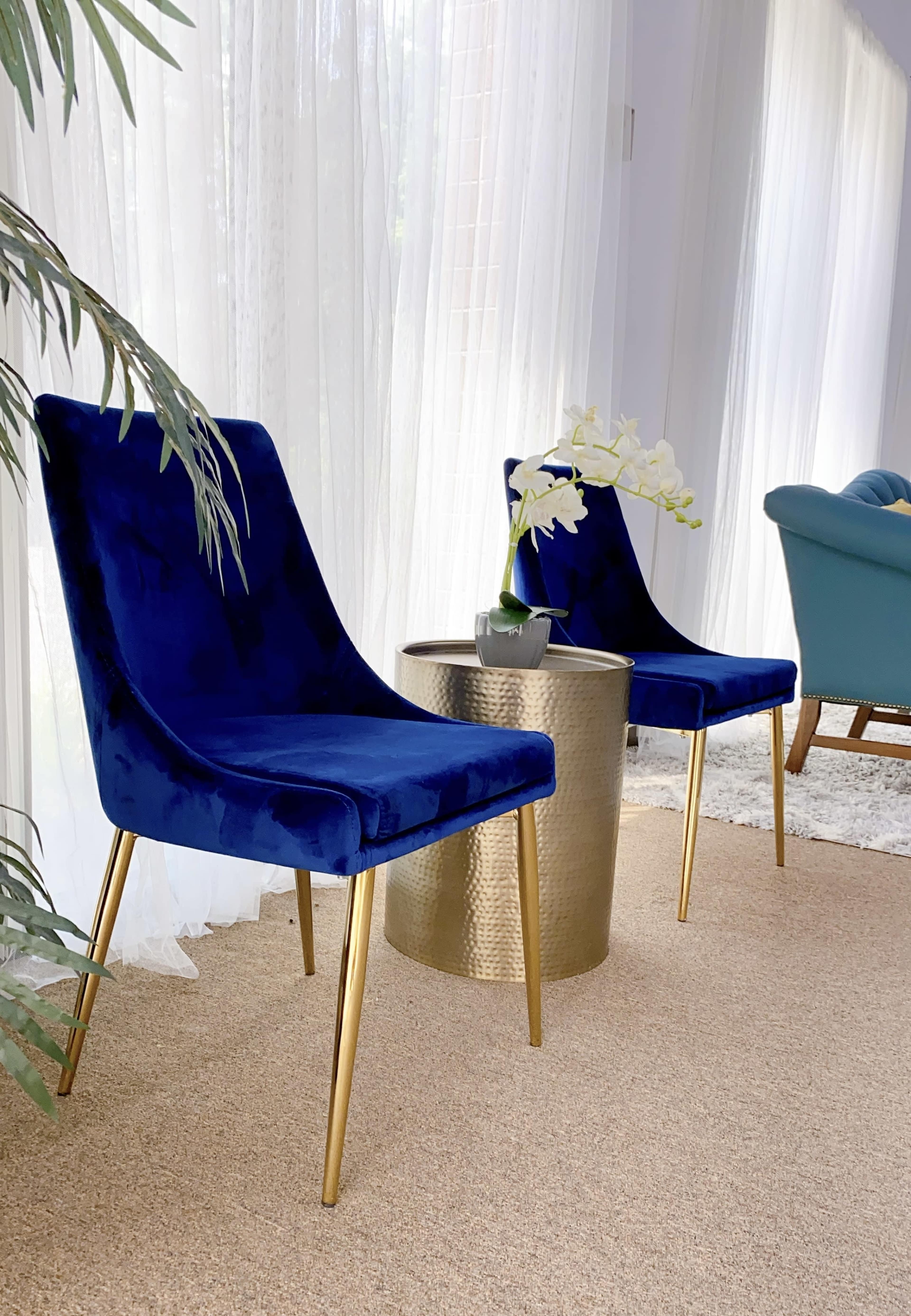 Two blue velvet chairs with gold legs sit next to a round, hammered gold table, adorned with a small potted orchid, in a light-filled room with sheer curtains.
