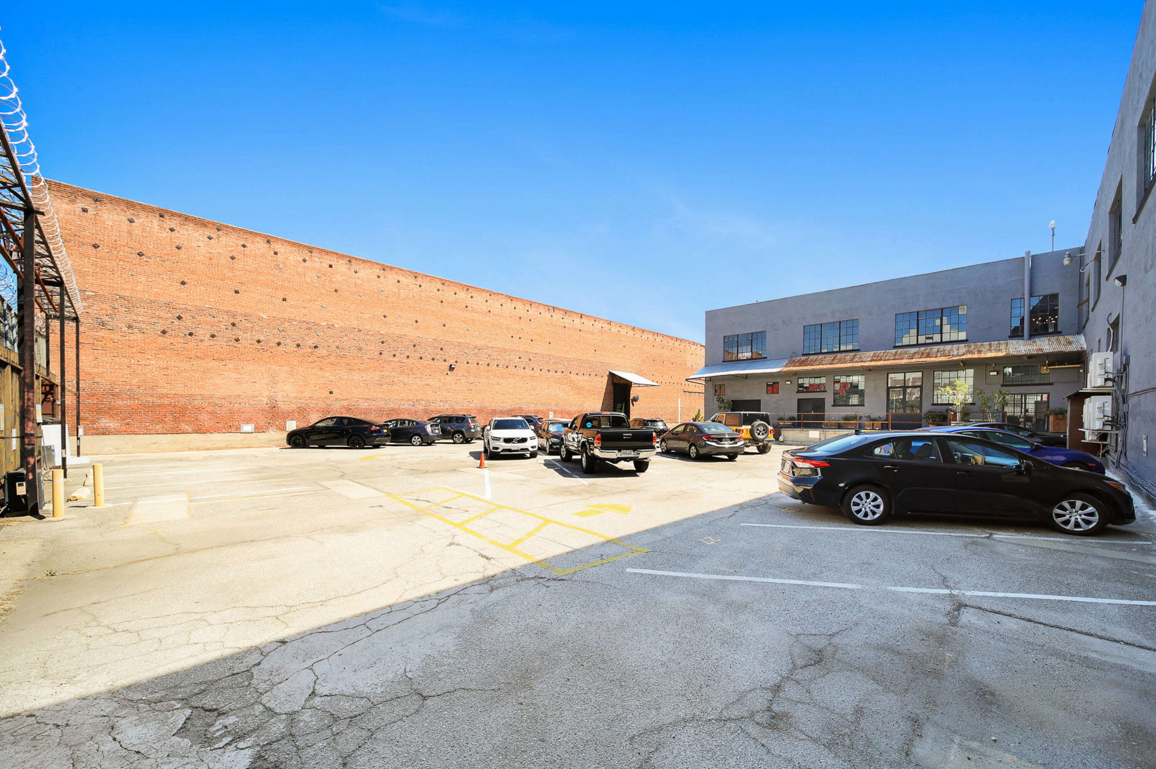 The image shows a parking area surrounded by brick and concrete buildings, with several parked cars and a clear blue sky above.