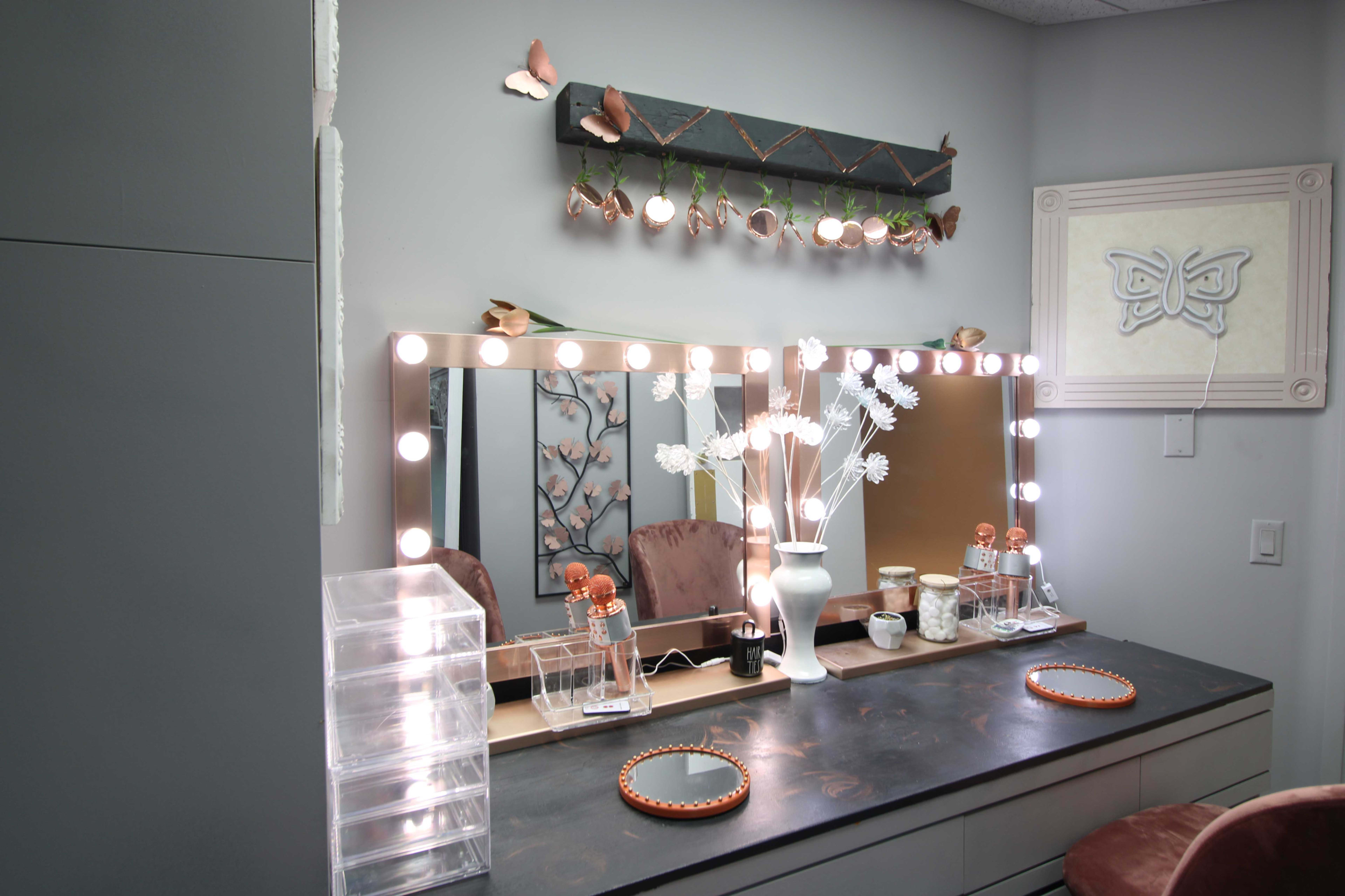 The image features a makeup vanity with a large mirror surrounded by lights, complemented by decorative items and a clean, organized surface.