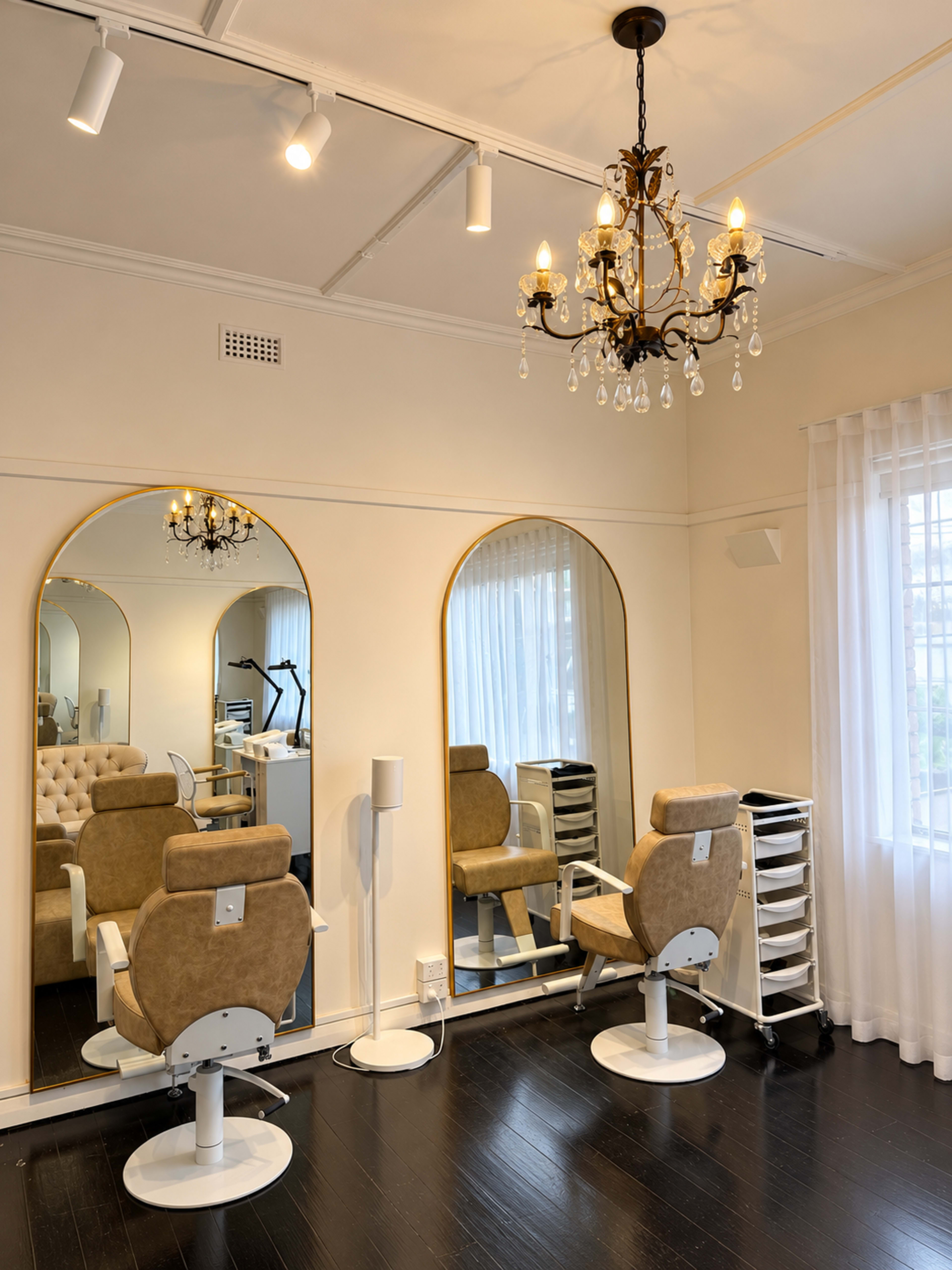 The image shows a bright, elegant salon interior featuring two styling chairs, large mirrors, a chandelier, and a light-colored decor with large windows.