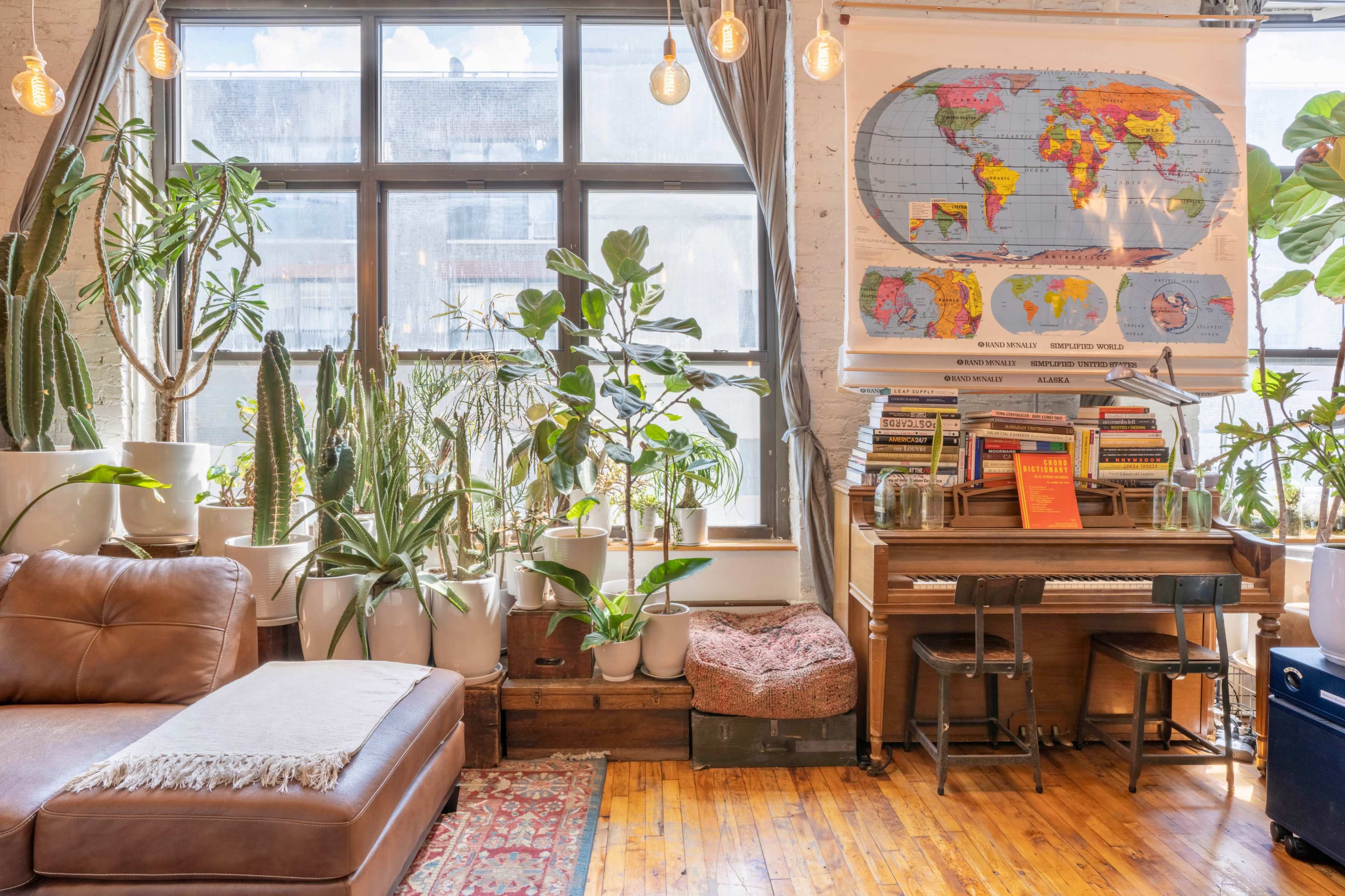 The scene features a cozy living space with a brown sofa, a vintage piano, a world map on the wall, and various plants in white pots near large windows.