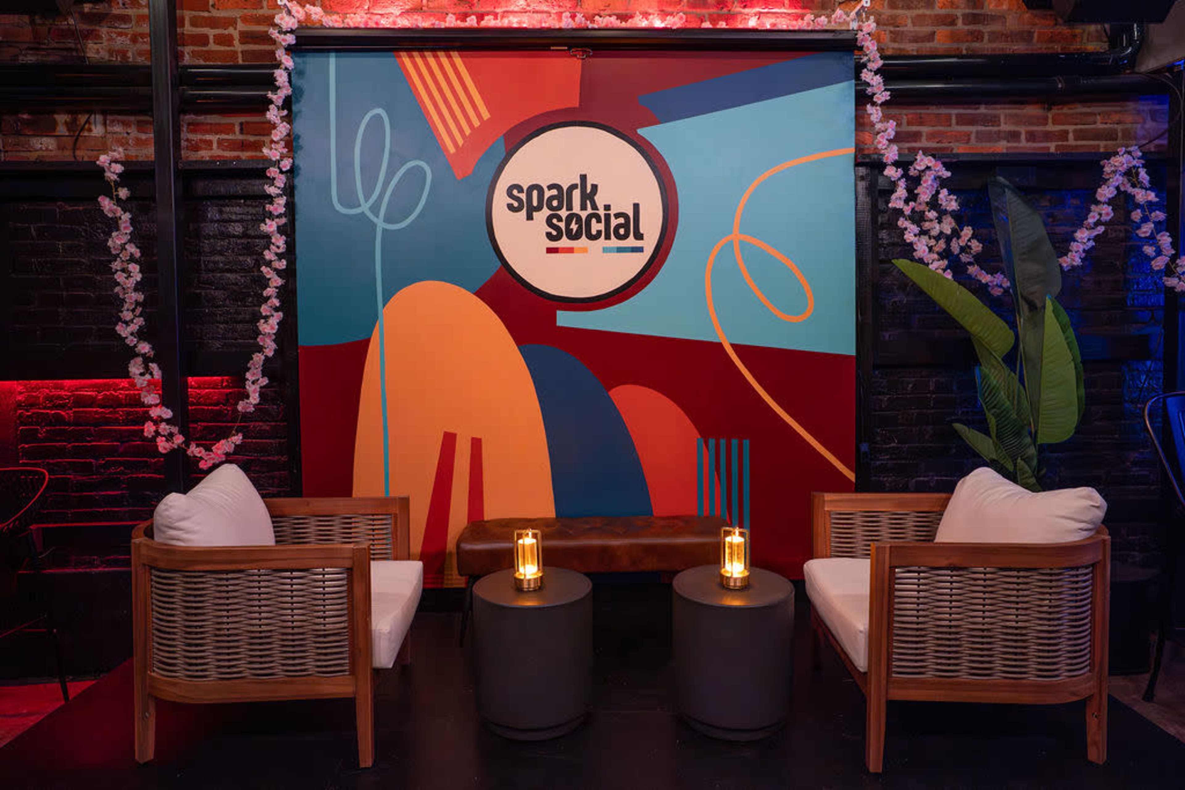 The image shows a cozy seating area with two chairs and small tables in front of a vibrant mural that features abstract shapes and the text "spark.social," surrounded by decorative plants and floral elements.
