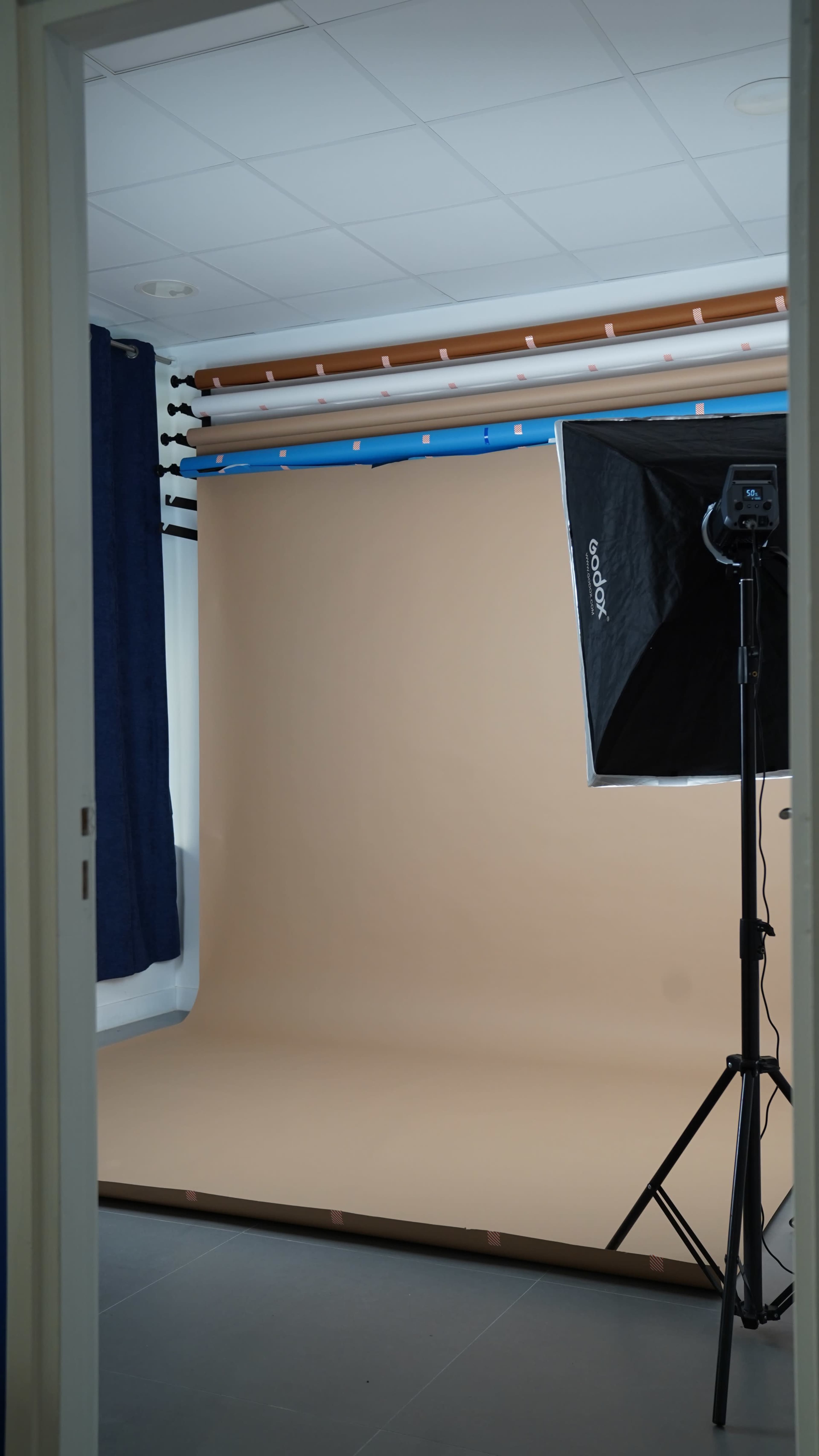 The image shows a photography studio with a beige backdrop and lighting equipment set up for a photoshoot.