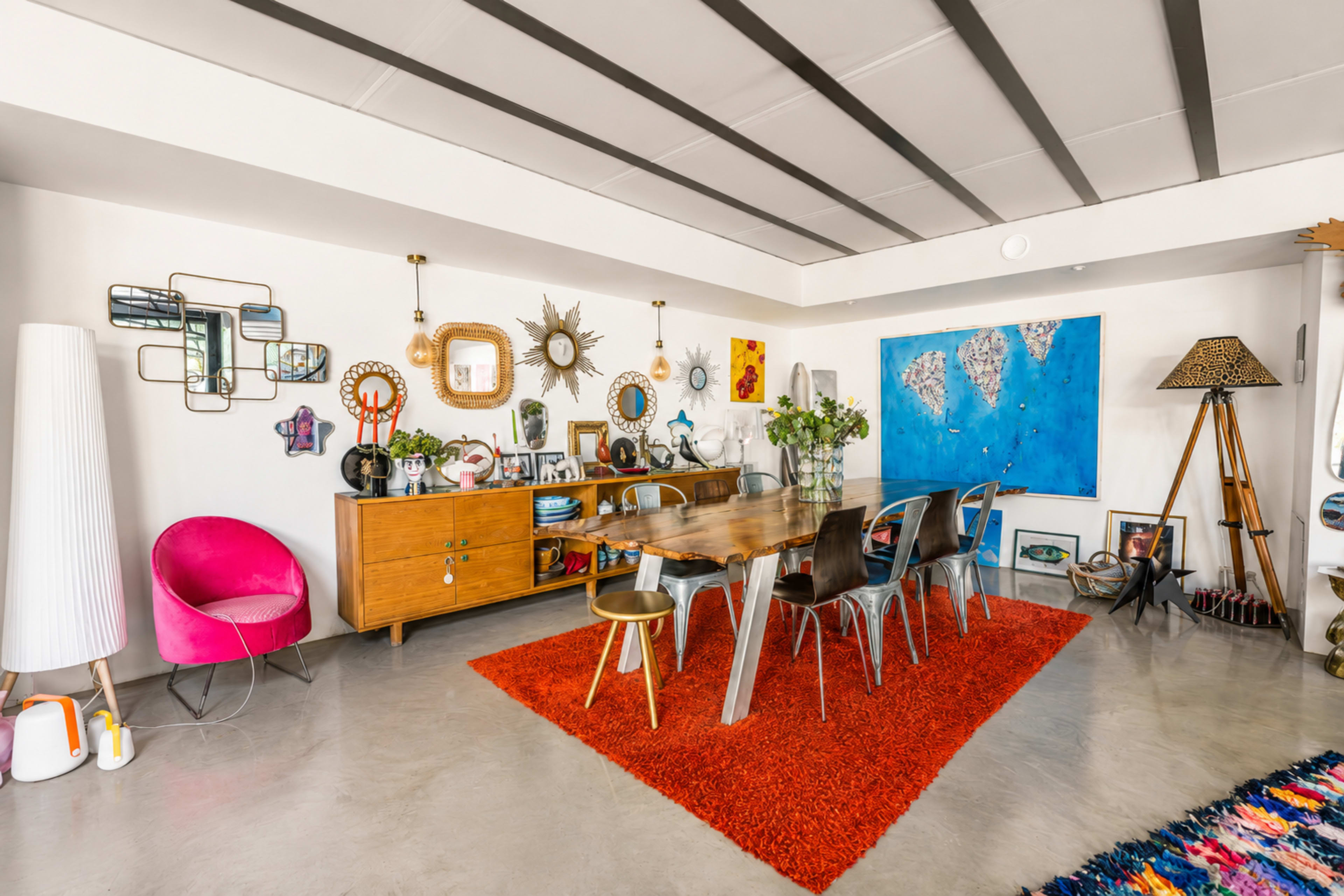 A dining area features a wooden table surrounded by metal and colorful chairs, with a red area rug and various decorative items on the walls and shelves.