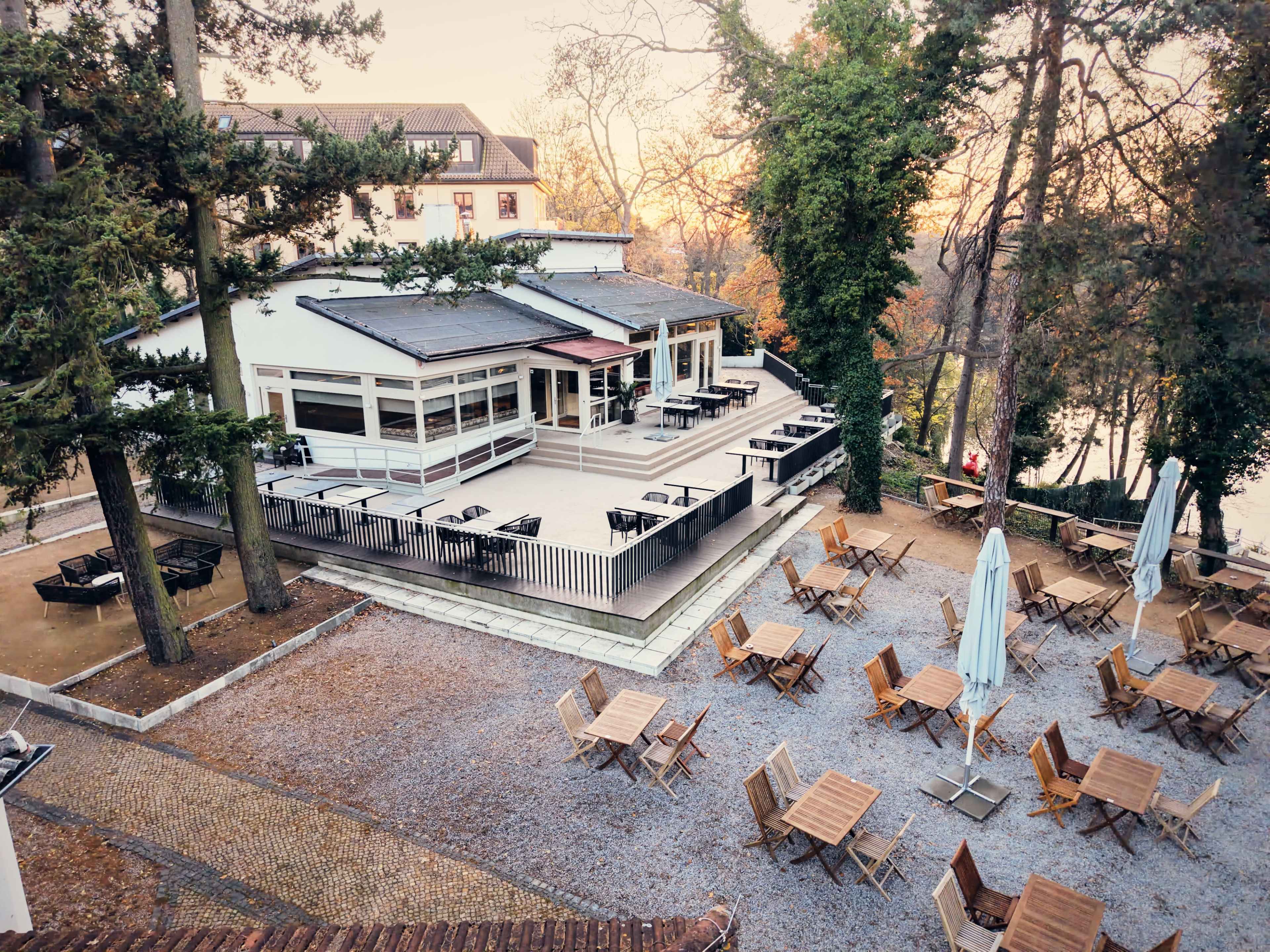The image shows a modern outdoor seating area of a restaurant surrounded by trees, featuring wooden tables and umbrellas.