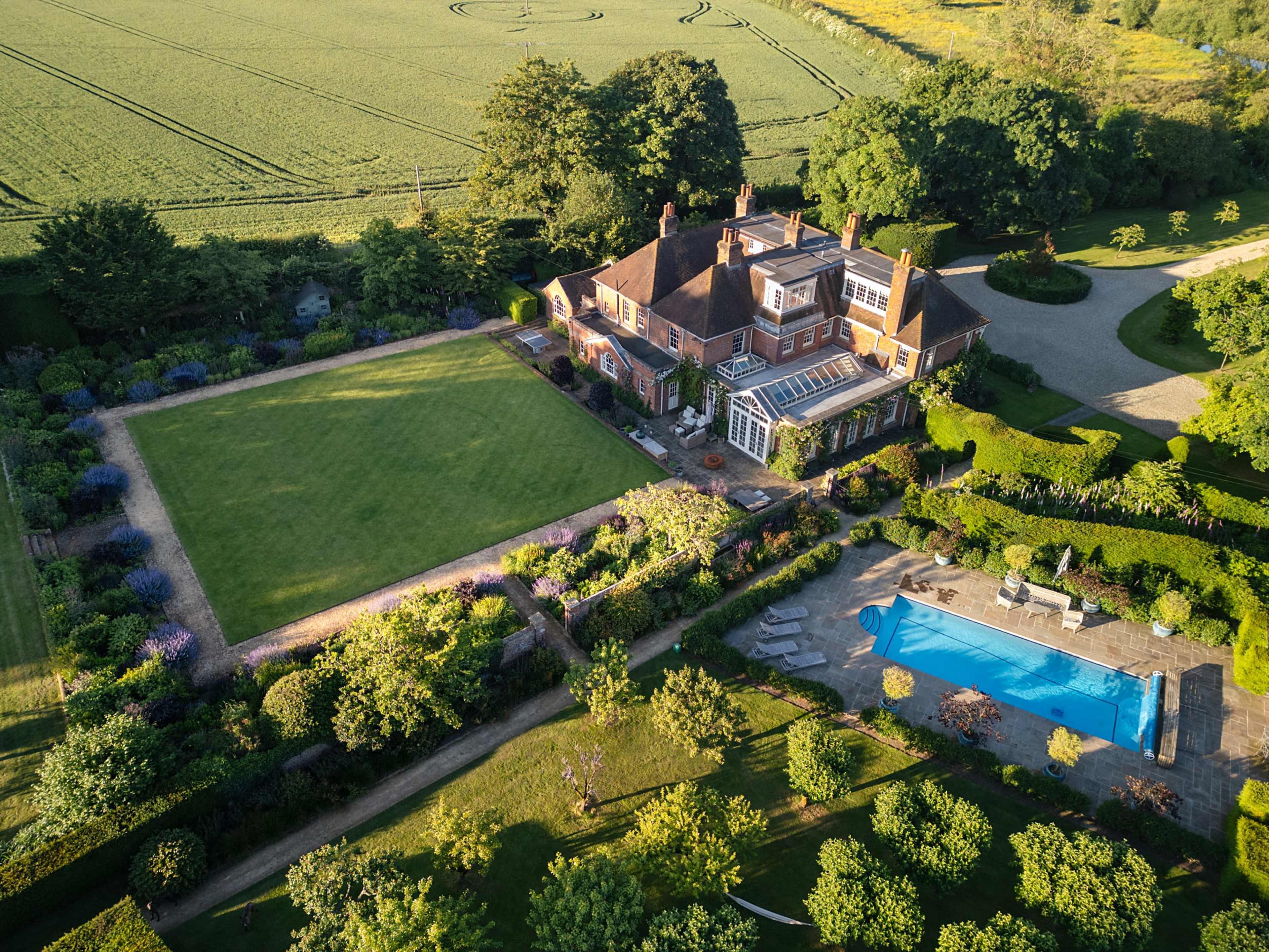 The image shows a large, elegant house surrounded by landscaped gardens, a swimming pool, and fields in the background.