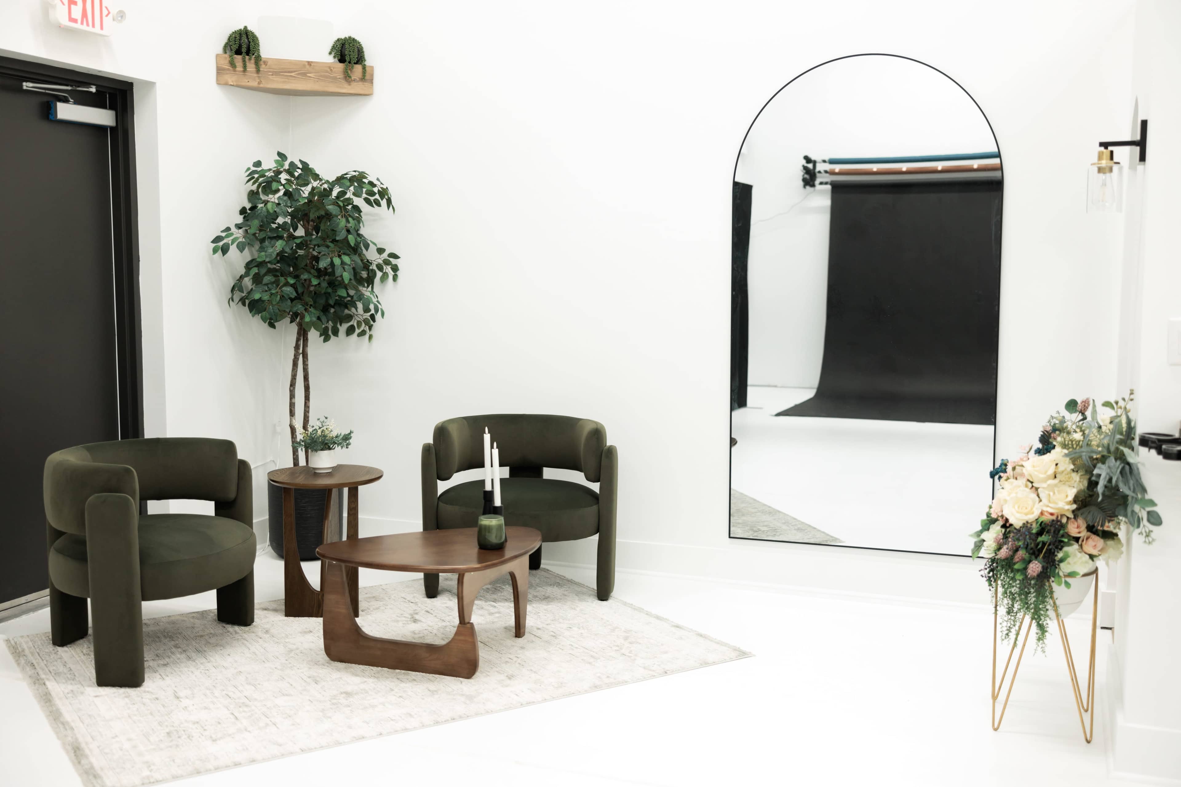 A small, stylish interior space features two green armchairs, a wooden coffee table, a large mirror, and a decorative plant against a white wall.