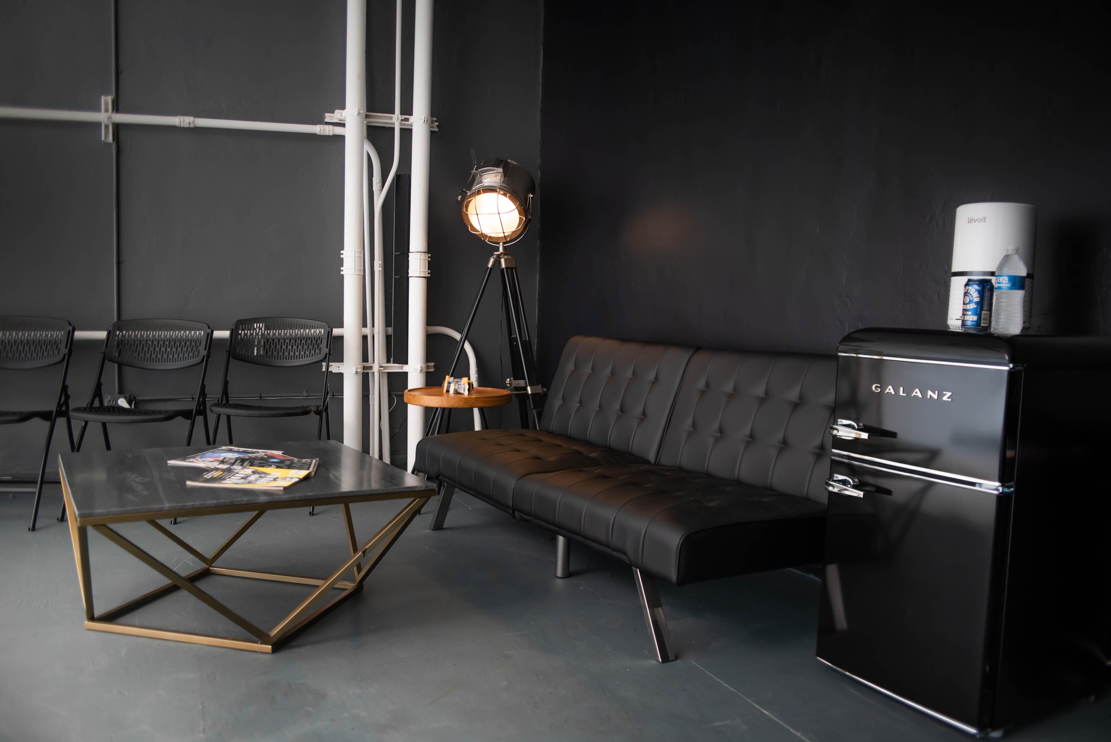 A modern lounge area featuring a black tufted sofa, a geometric coffee table, a vintage-style floor lamp, a small wooden side table, and a black refrigerator, all set against a dark wall with exposed pipes.